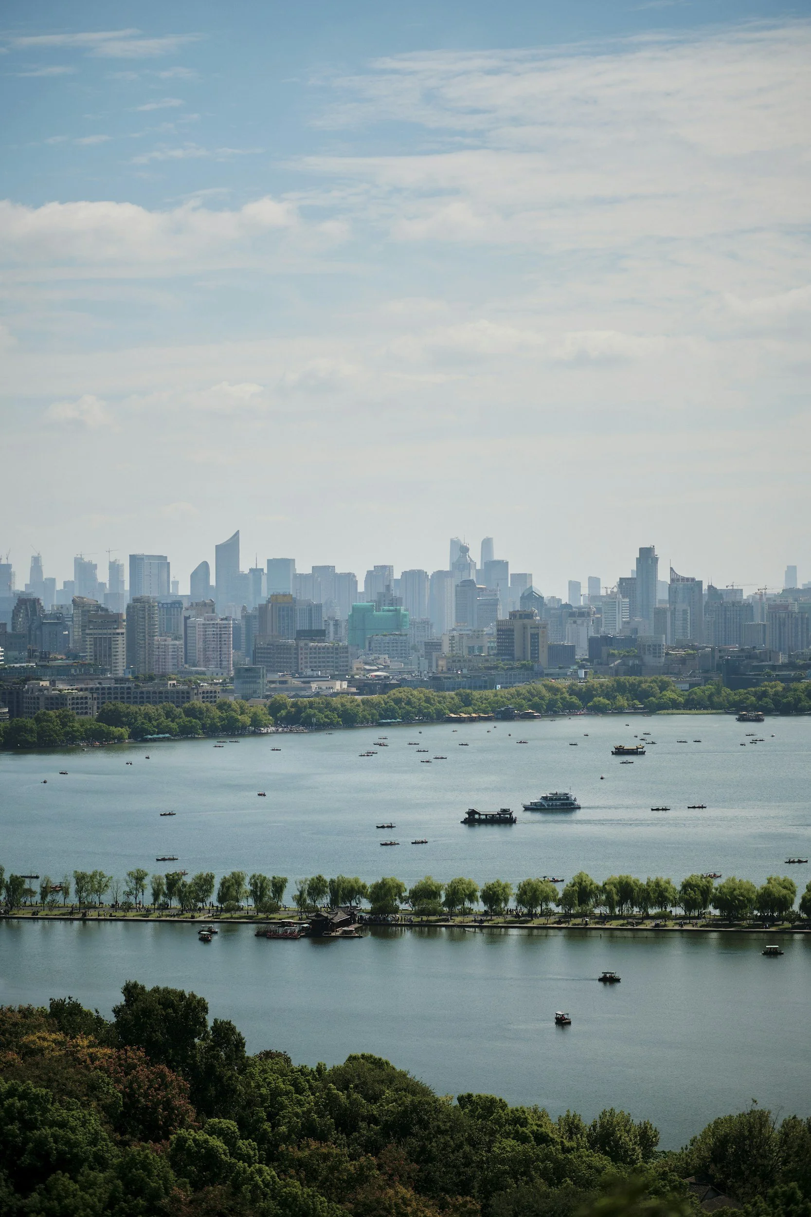 View of a city skyline across a body of water with boats, surrounded by trees and greenery.
