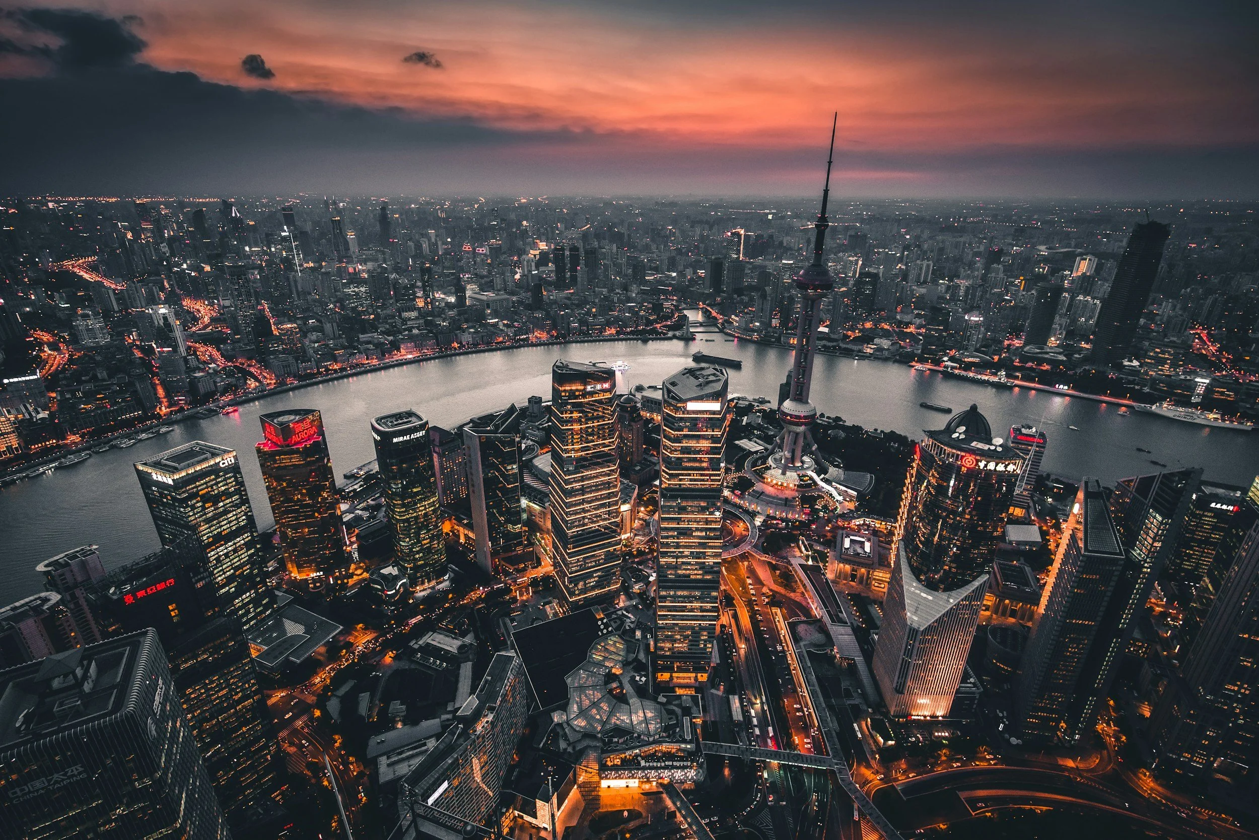 Aerial view of a city skyline at dusk, featuring tall skyscrapers, a river, and a large TV tower in the center with a sunset sky in the background.