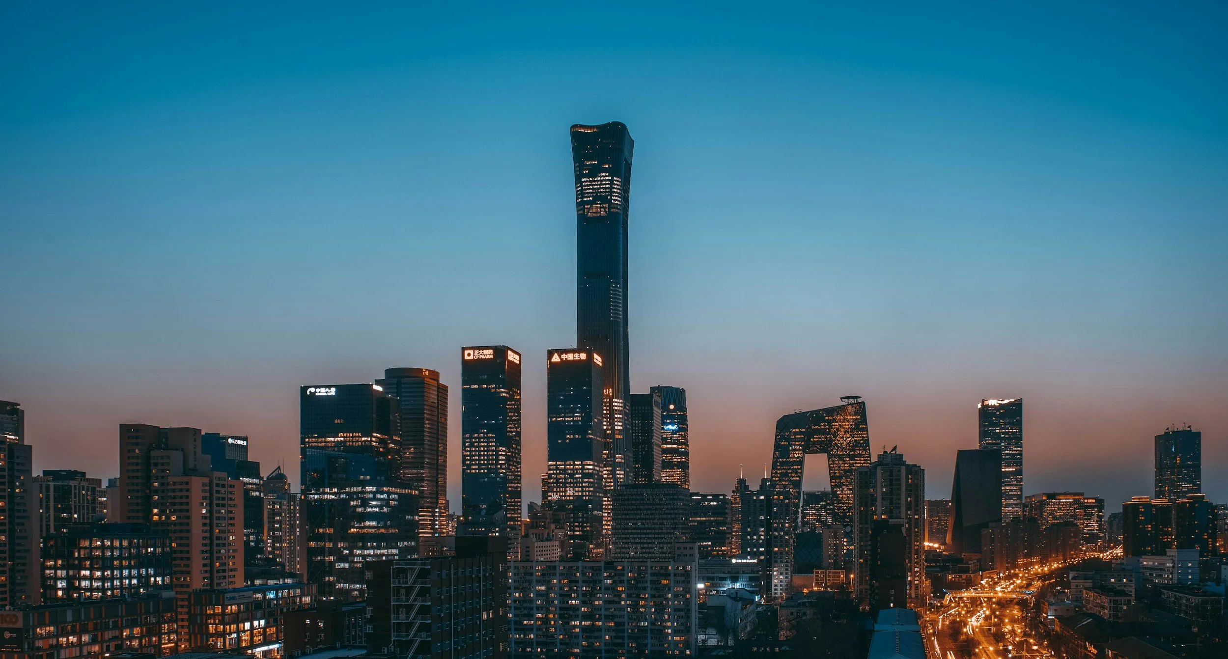 City skyline at dusk featuring tall skyscrapers, including the prominent China Zun Tower, illuminated windows, and streetlights.