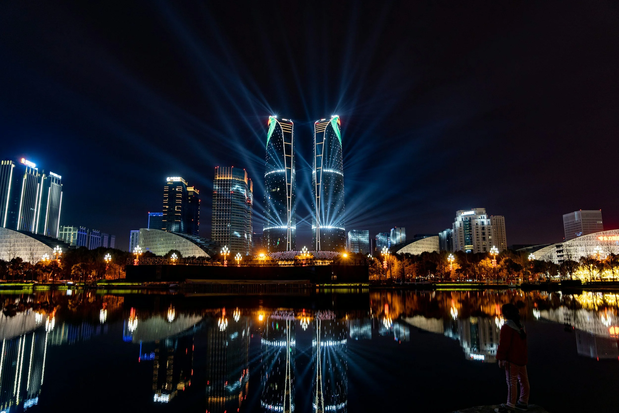 Nighttime cityscape with illuminated skyscrapers and a light show on two tall buildings, reflected in a body of water, with a person standing near the water's edge.