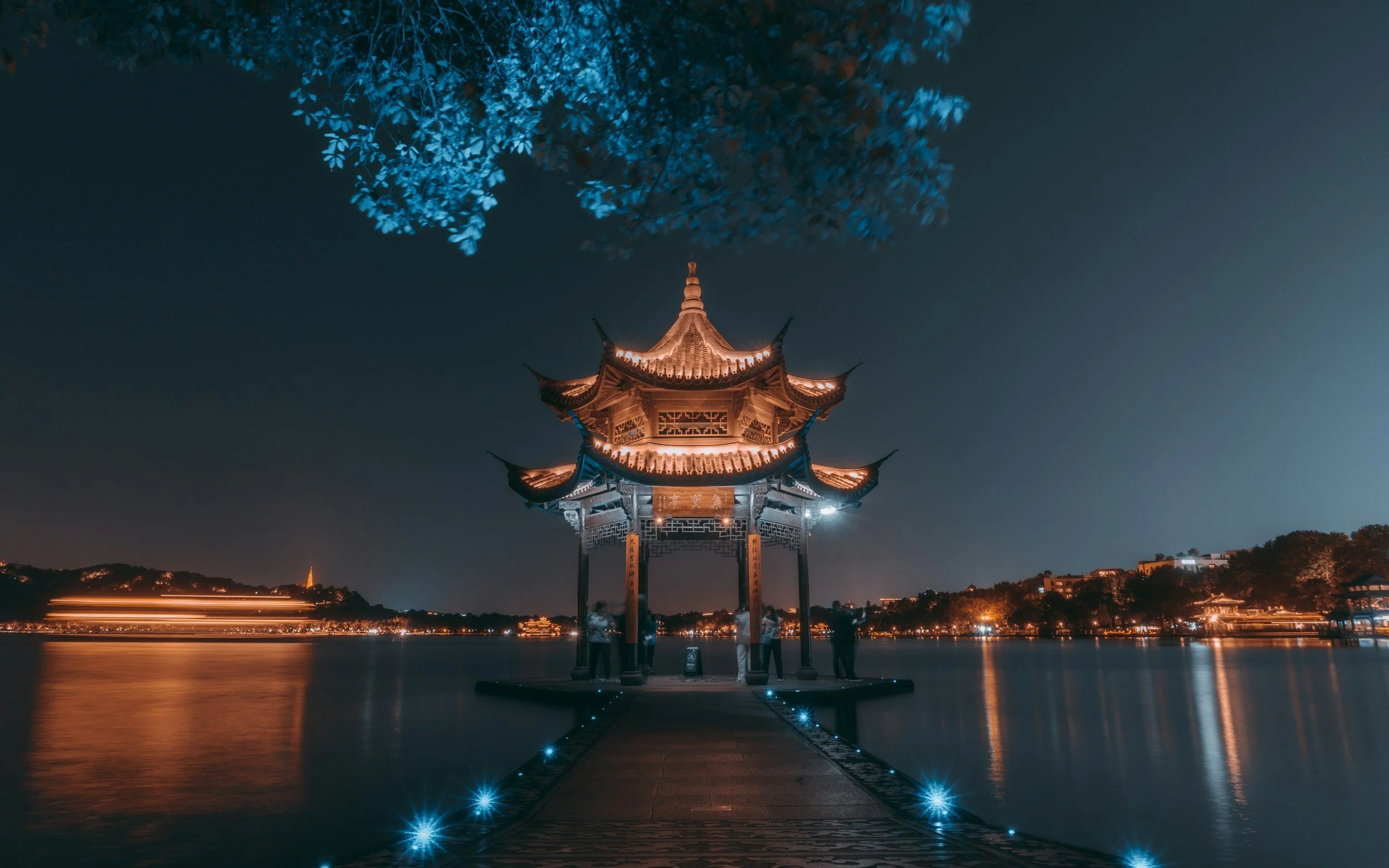 Night view of a traditional Chinese pagoda by a lake, illuminated with warm lights, with trees and city lights in the background.