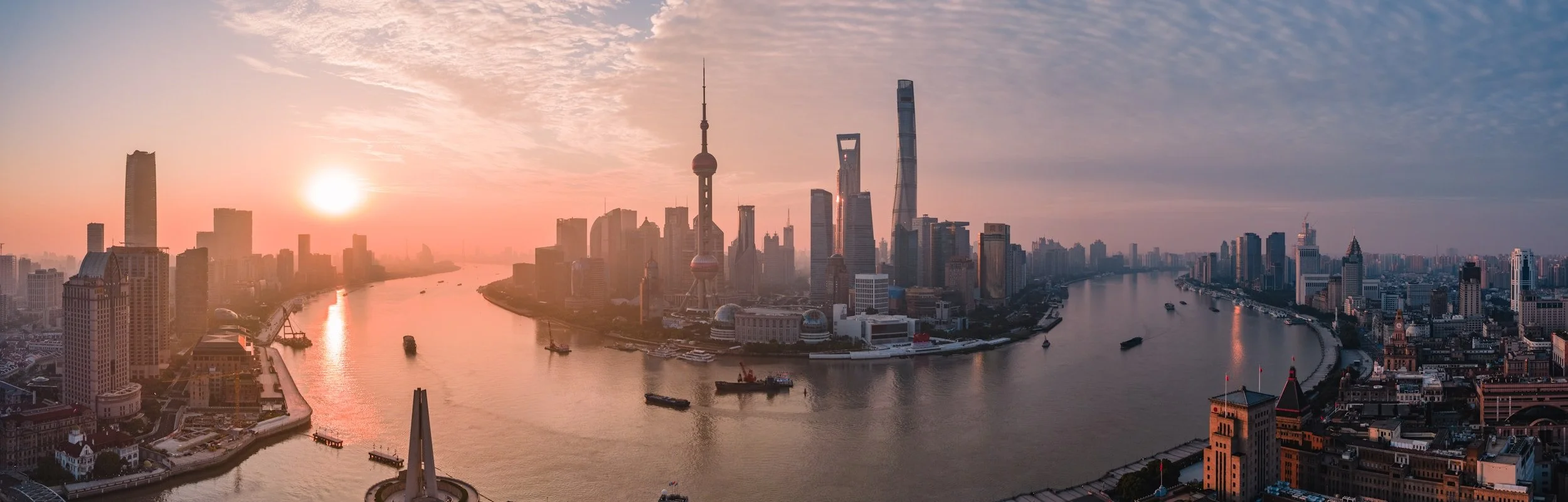 Panoramic view of Shanghai skyline at sunset, with tall skyscrapers, the Huangpu River, and boats on the water.