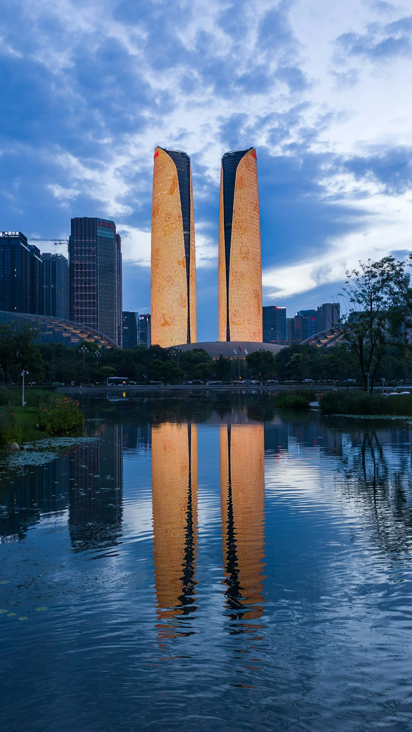 Nighttime cityscape with illuminated skyscrapers and a light show on two tall buildings, reflected in a body of water, with a person standing near the water's edge.