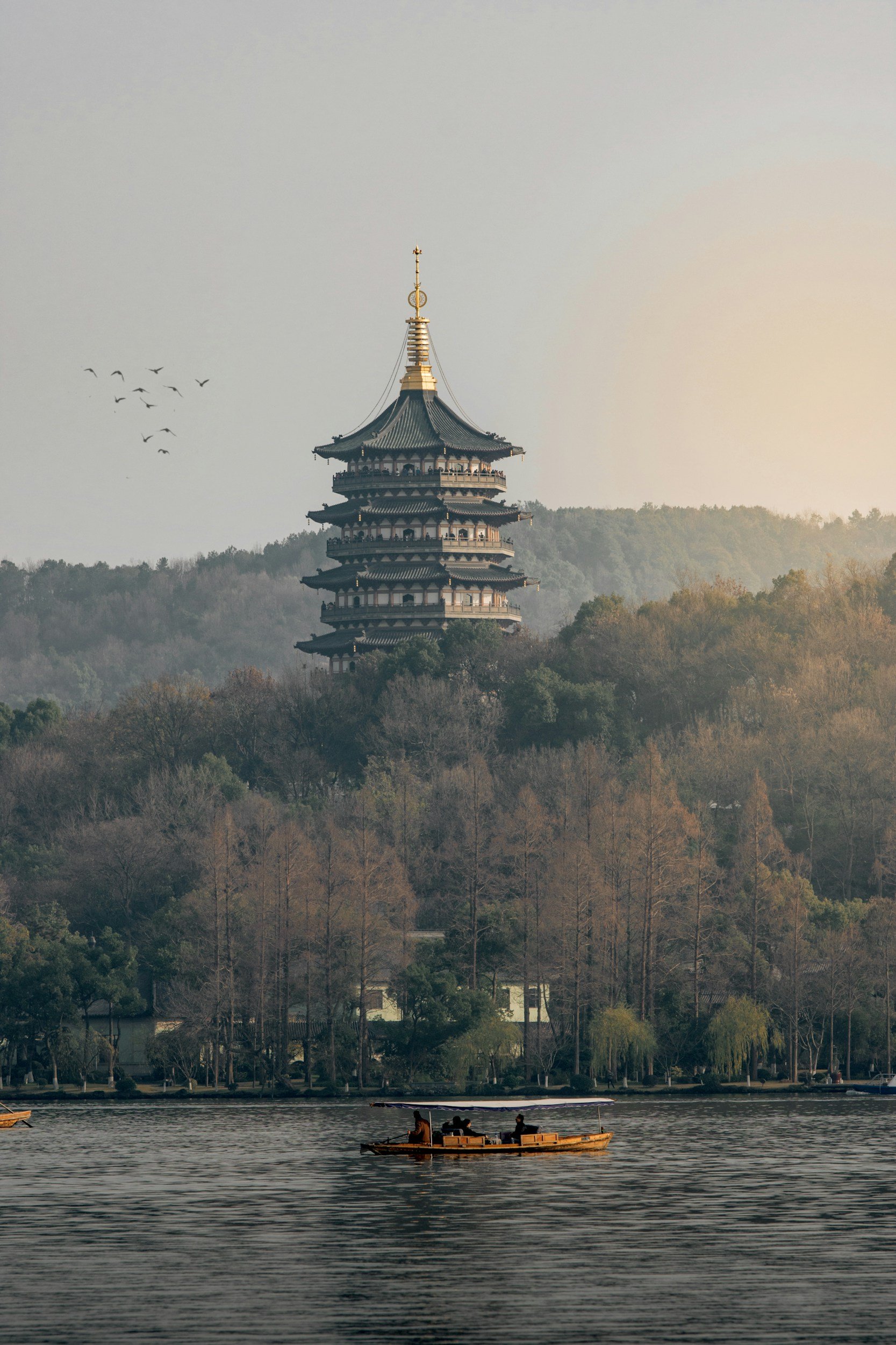 Night view of a traditional Chinese pagoda by a lake, illuminated with warm lights, with trees and city lights in the background.