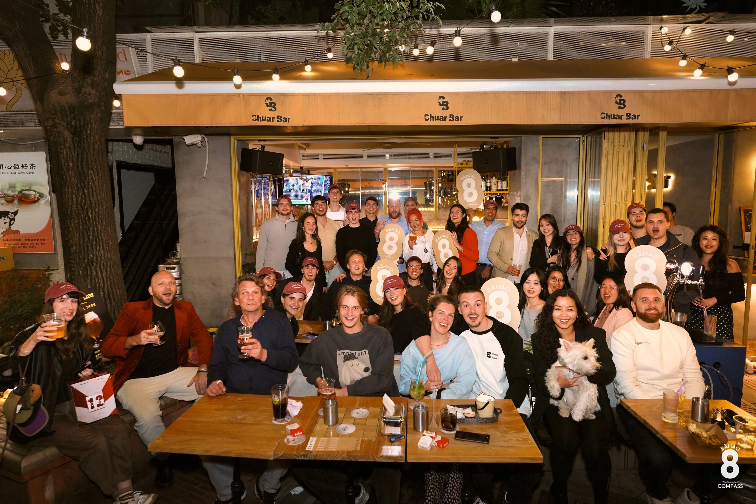 Group of people gathered at a celebration in a restaurant or bar, some holding drinks and signs with the number 8, with a mix of standing and seated individuals, a large dog, and decorative lighting.