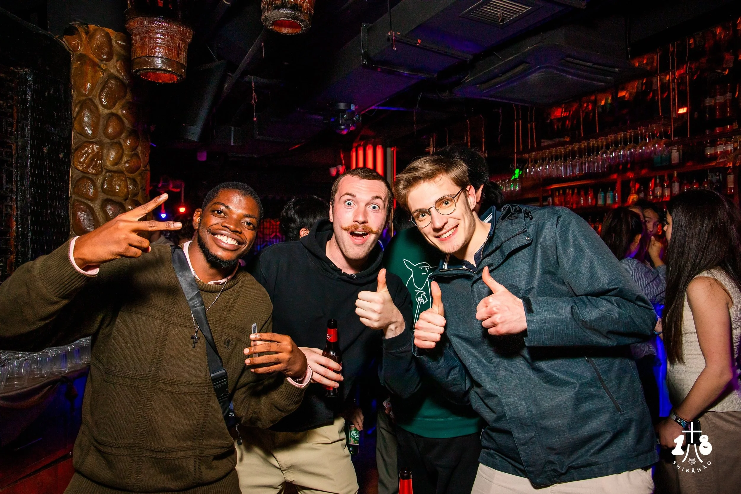 Three men at a party or nightclub, smiling and making hand gestures, with a bar and other people in the background.