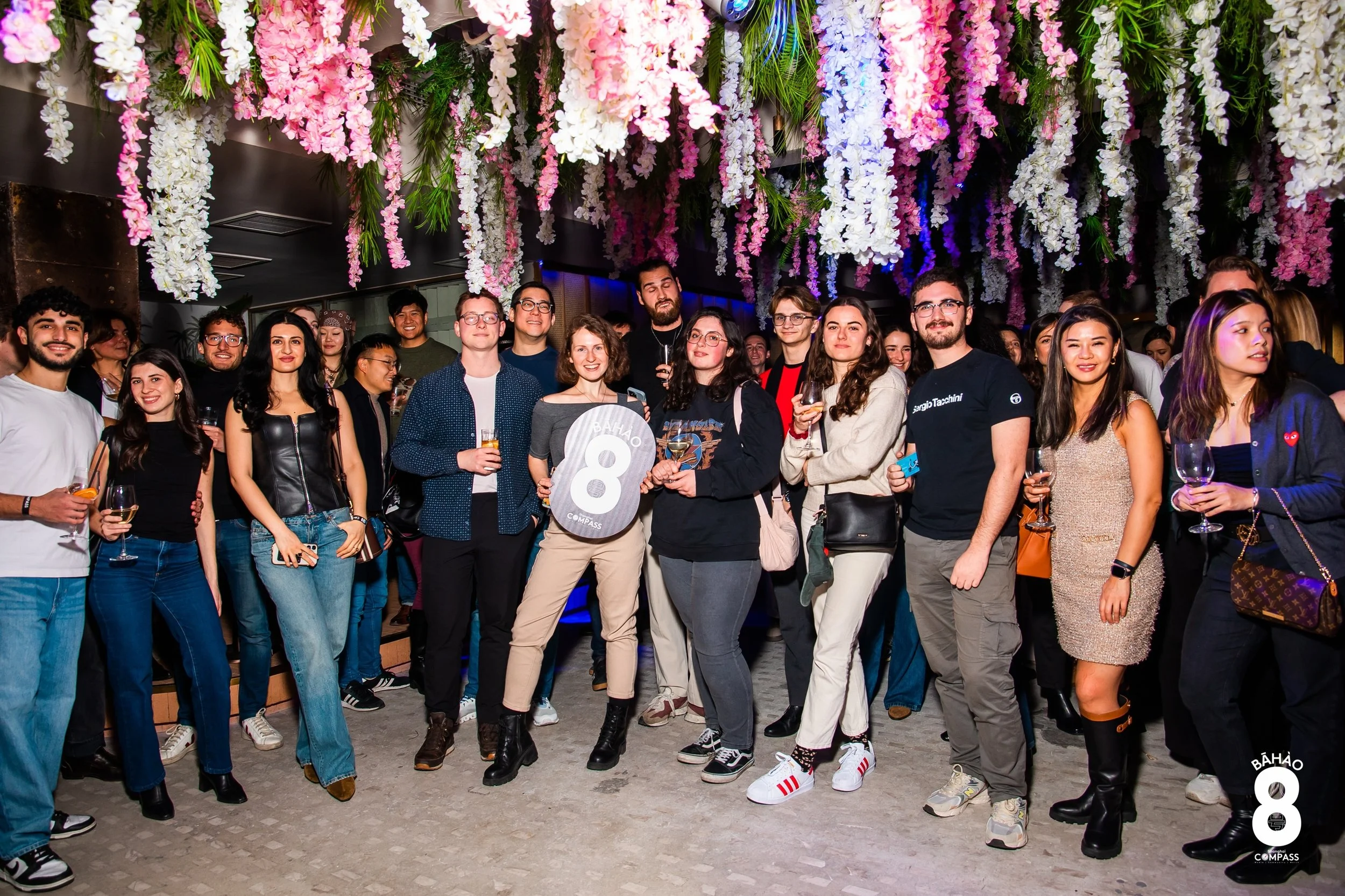 A group of people at a celebration or party, standing together under hanging pink and white floral decorations, with some holding drinks and smiling at the camera.