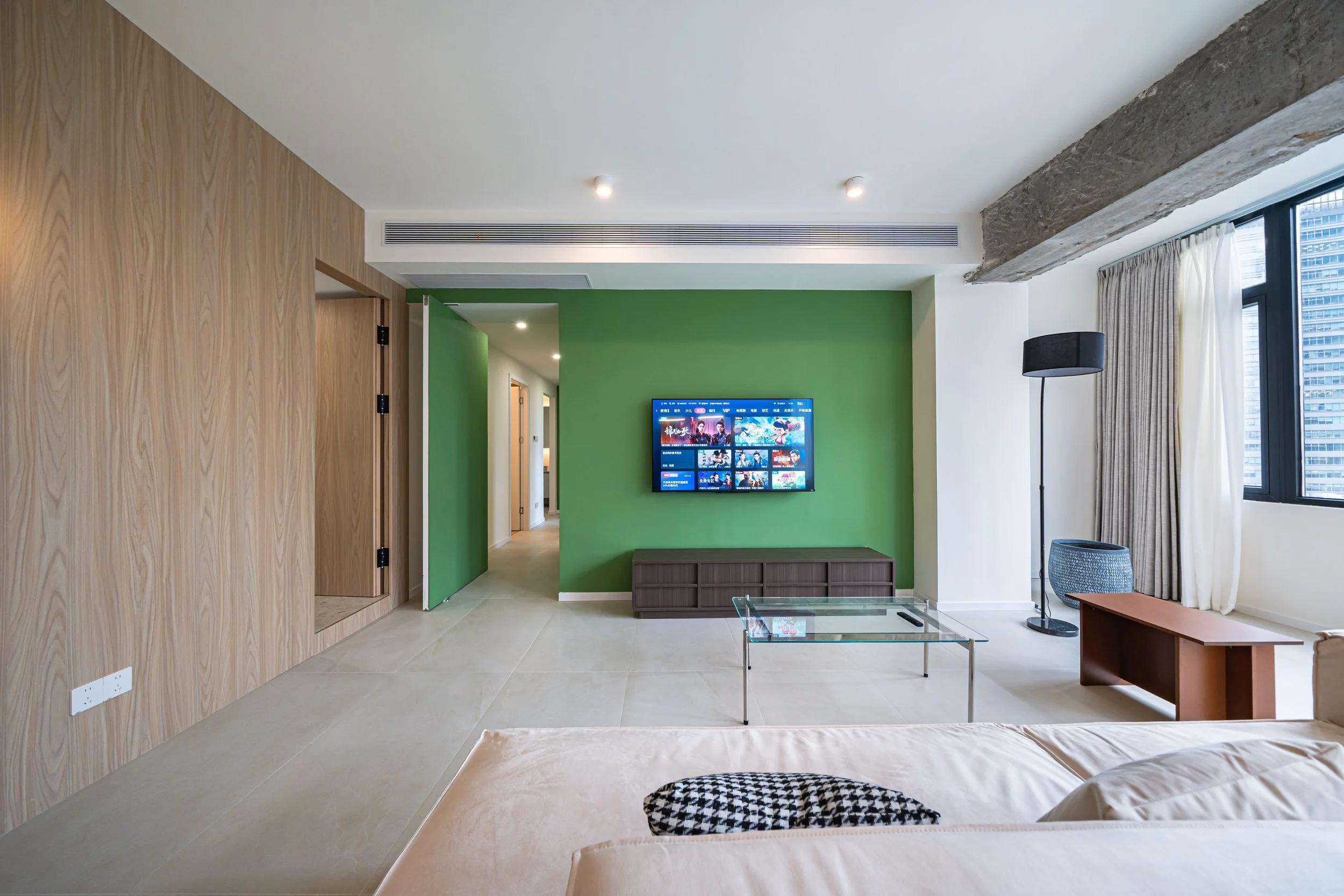 Modern living room with green accent wall, wall-mounted TV, glass coffee table, beige sofa, wood paneling, large window with curtains, floor lamp, and city view outside.