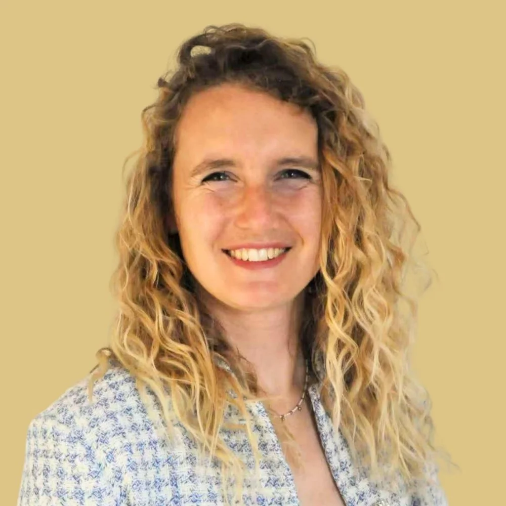 A woman with curly blonde hair smiling in front of a beige background.