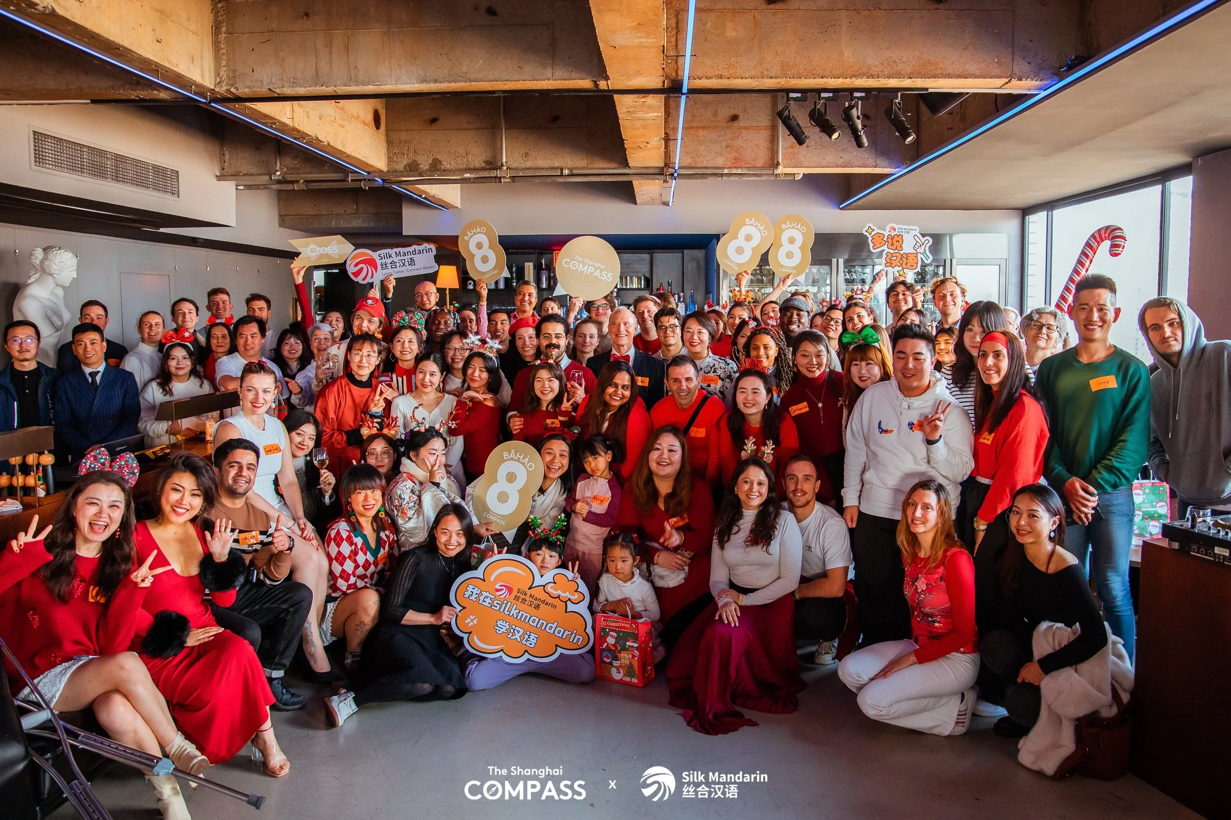 A large group of people gathered for a photo indoors, celebrating with Christmas decorations, some wearing festive clothing, and holding signs with logos and messages.