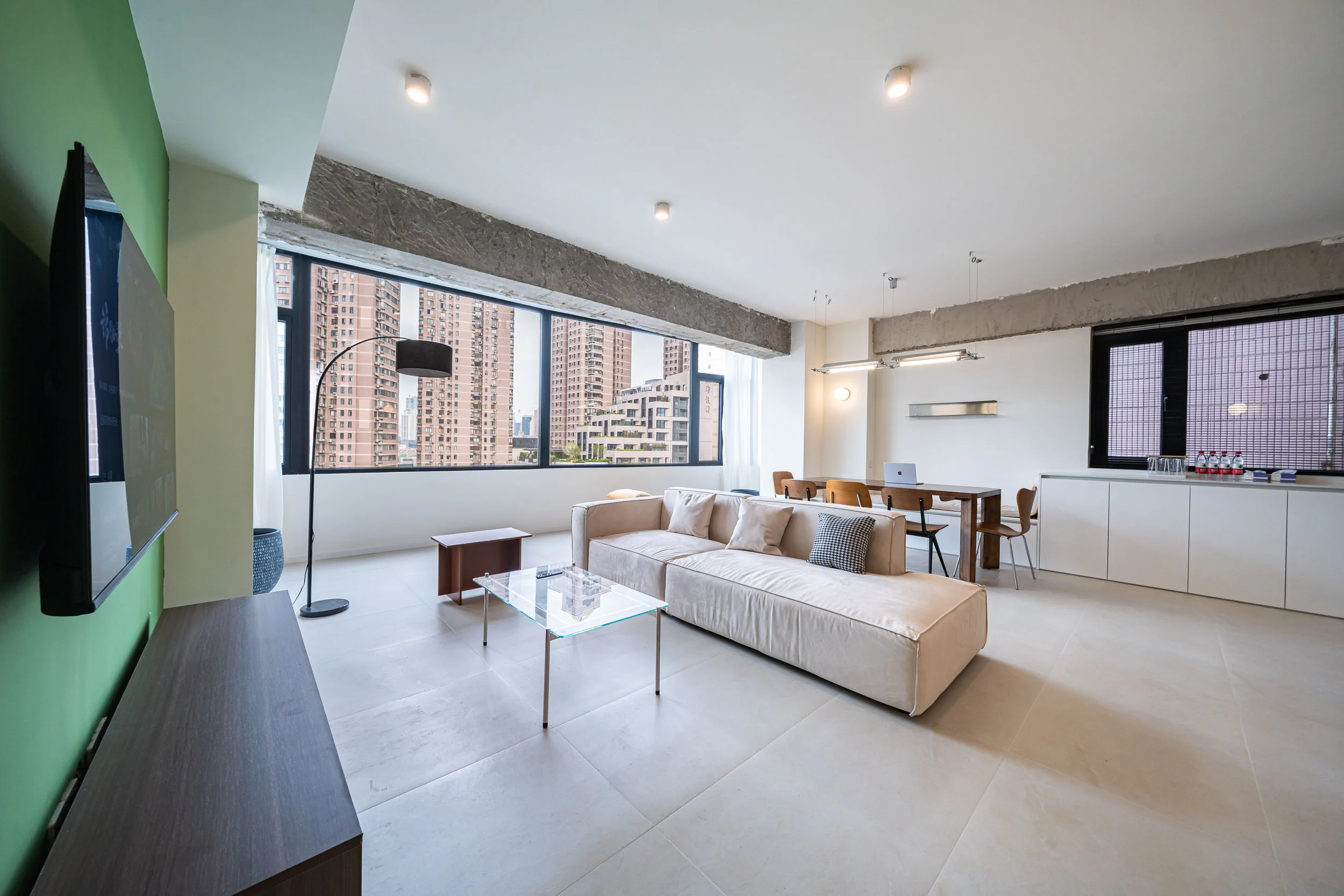 Modern city apartment living room with large windows, beige sofa, glass coffee table, and kitchen area with bottles on counter.