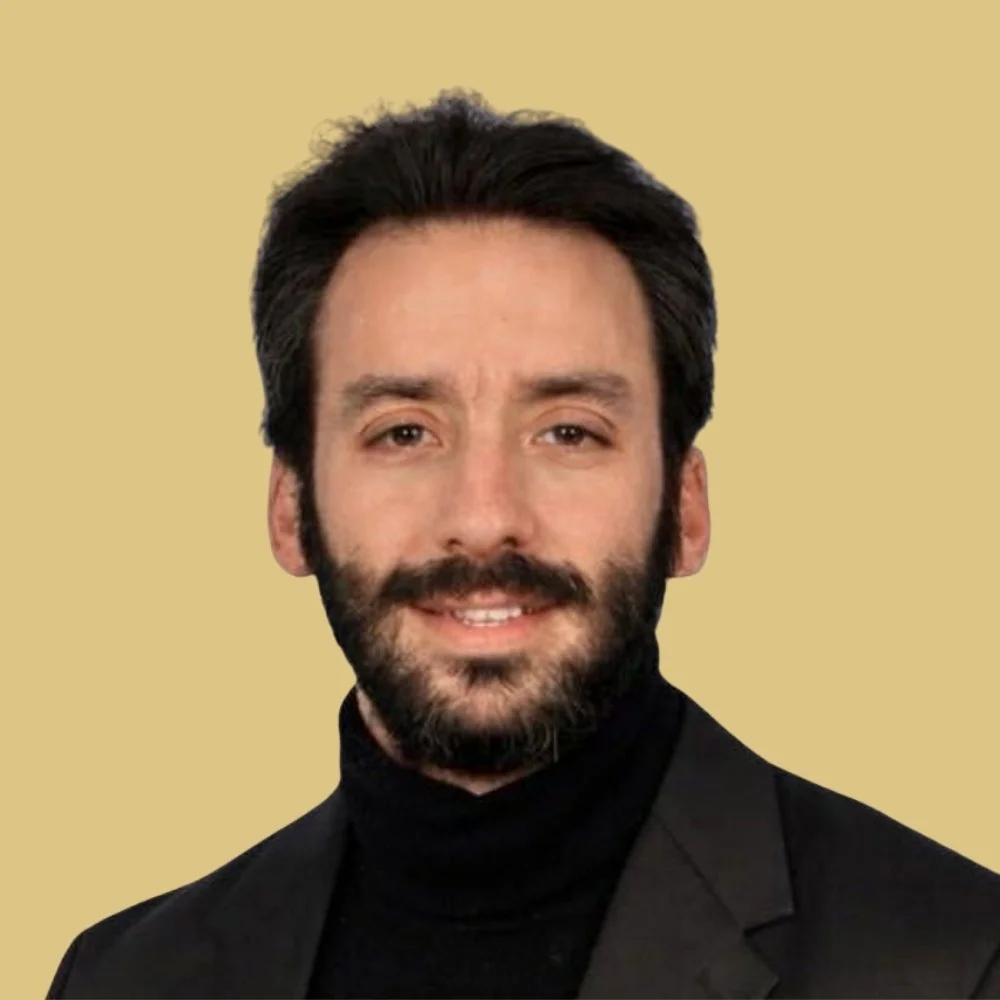 A man with dark hair, a beard, and mustache, wearing a black turtleneck and blazer, smiling slightly against a beige background.