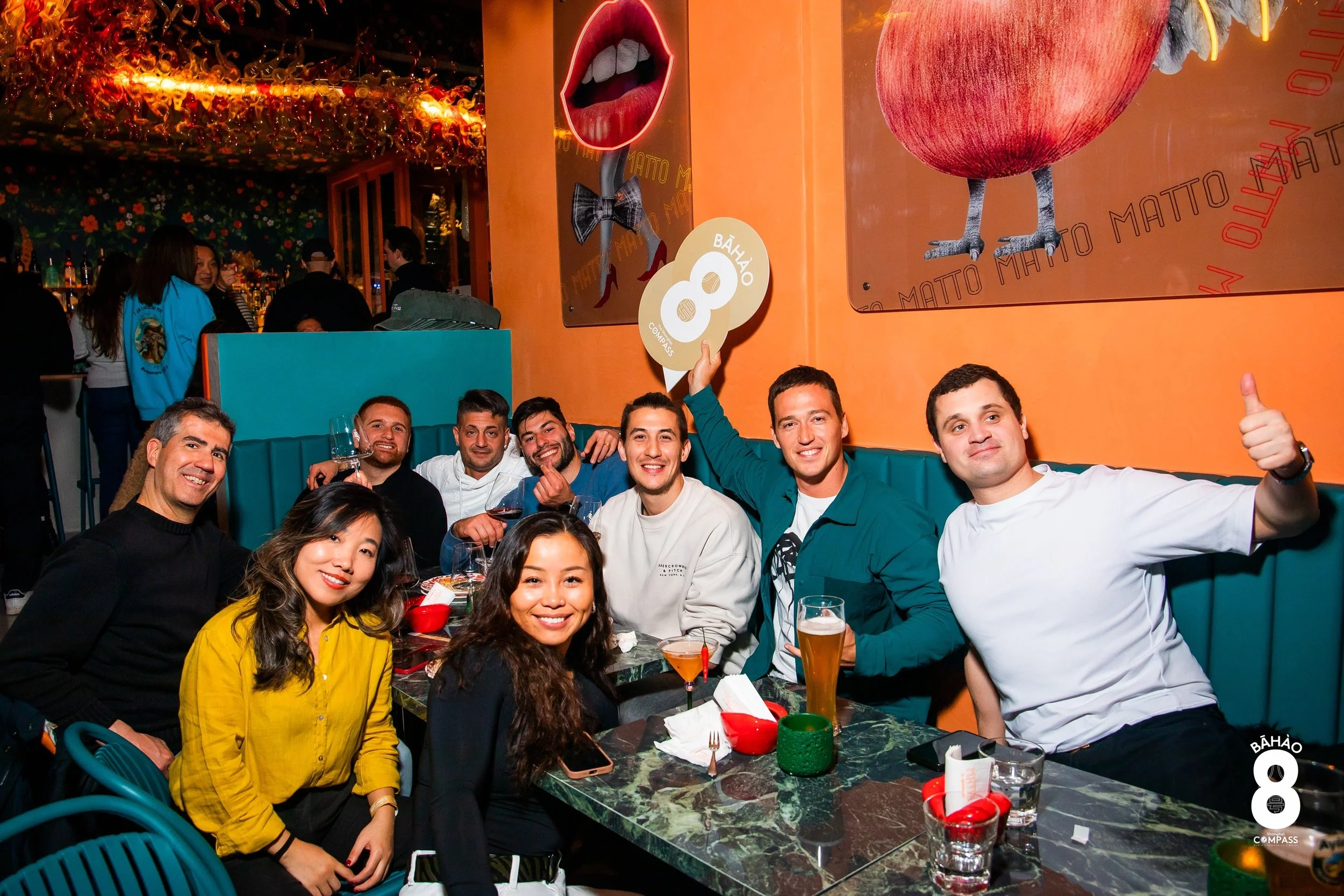 A group of nine diverse people gathered around a table at a lively restaurant or bar, smiling and enjoying drinks, with colorful wall art and decorations in the background.
