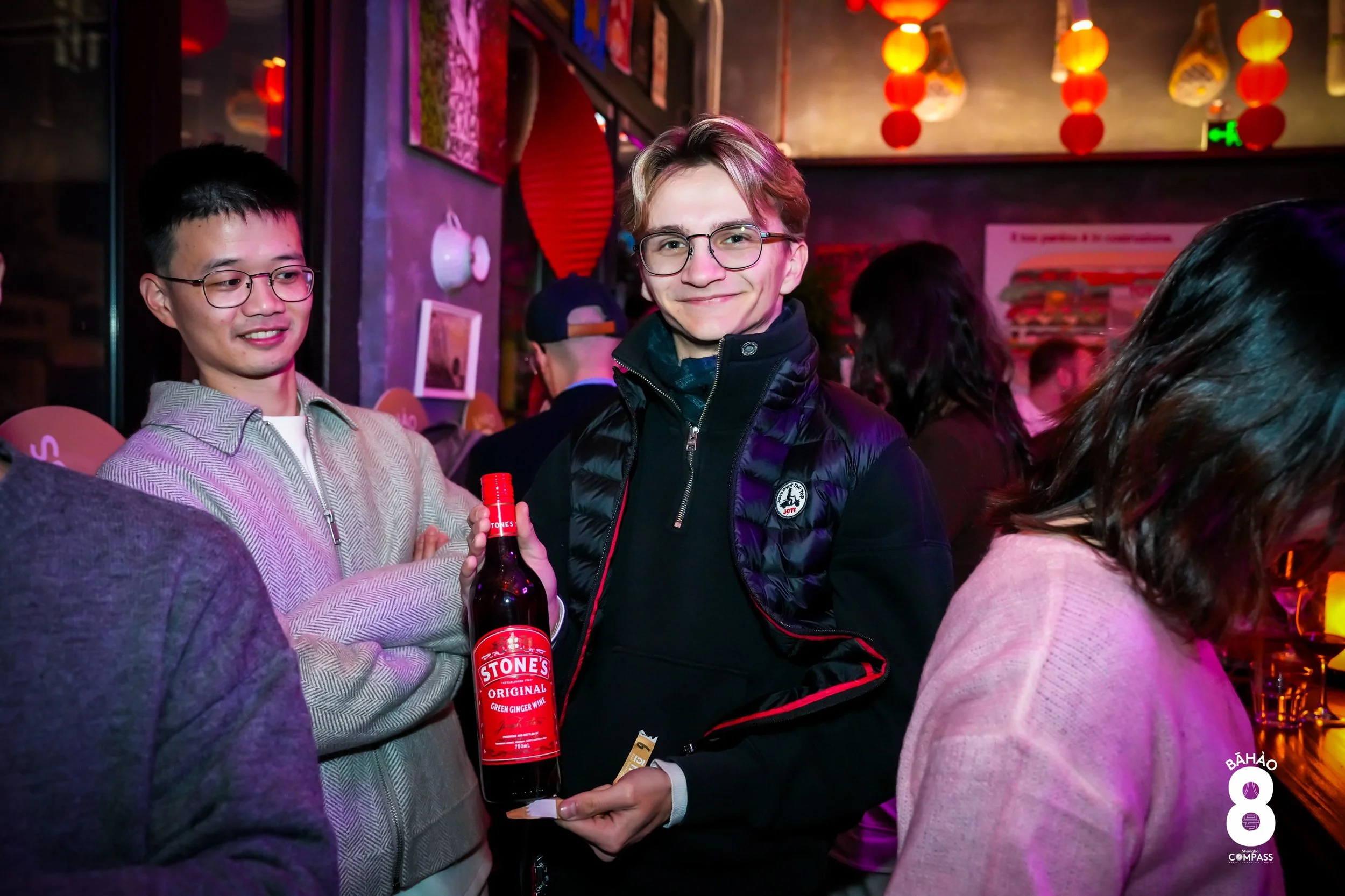 Young man in glasses holding a bottle of Stone's Original Green Ginger Wine at a lively indoor social gathering with colorful lanterns and other people in the background.