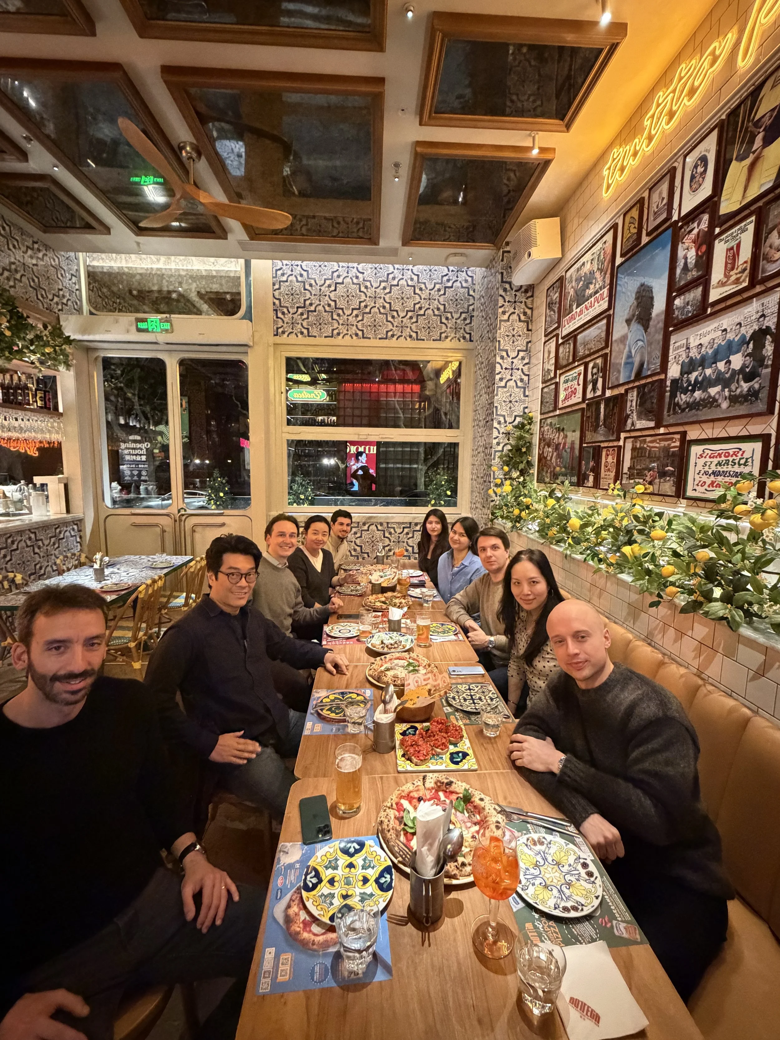 A group of twelve diverse adults sitting at a long table in a restaurant. The table has food, drinks, and napkins. The interior features decorative tiled walls, framed photos, and a lemon and greenery display on the wall.