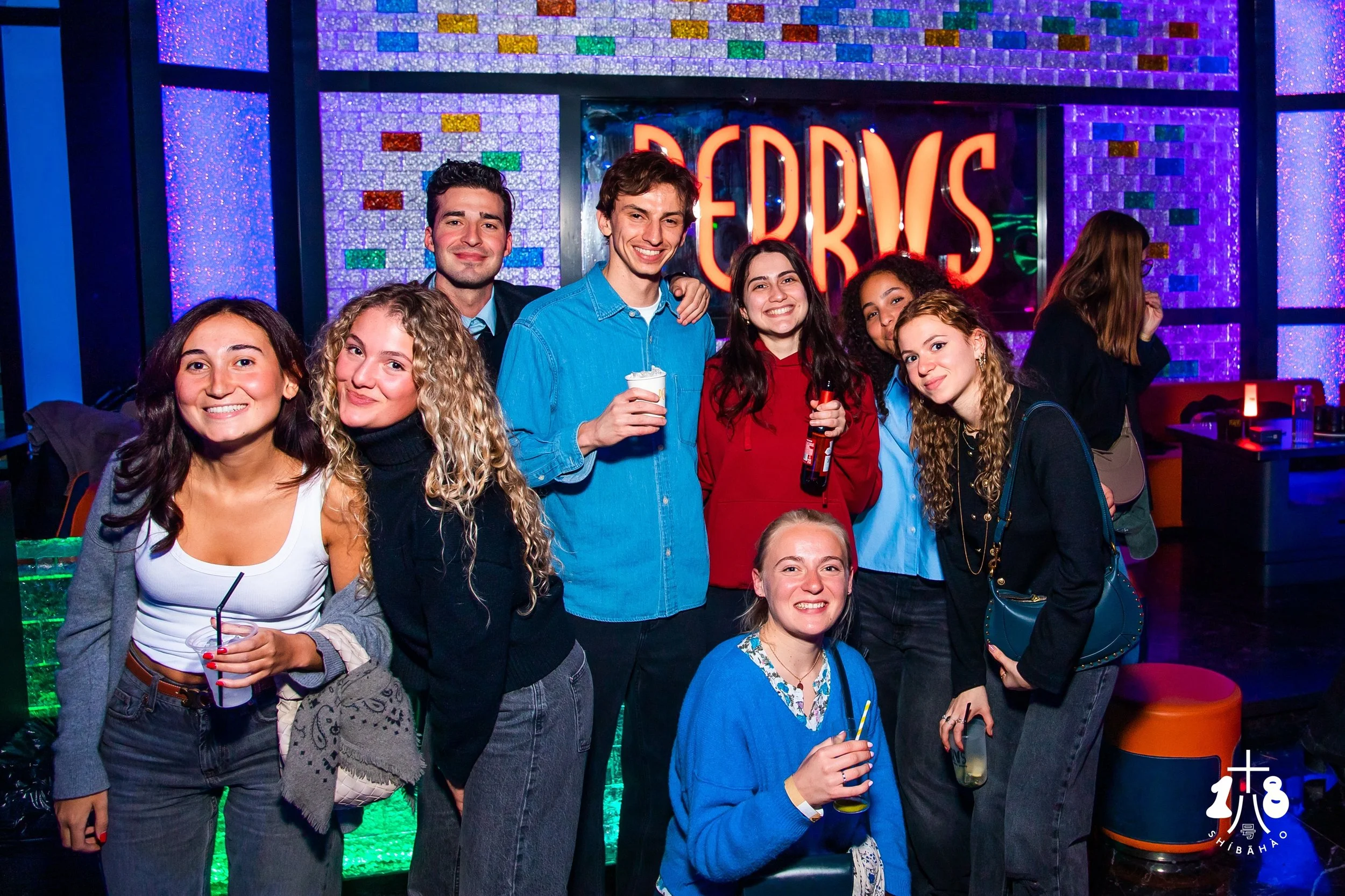 Group of eight young adults smiling and posing for a photo at a colorful nightclub or bar, with a neon sign in the background that reads 'DRINKS' and a vibrant, glittering wall behind them.