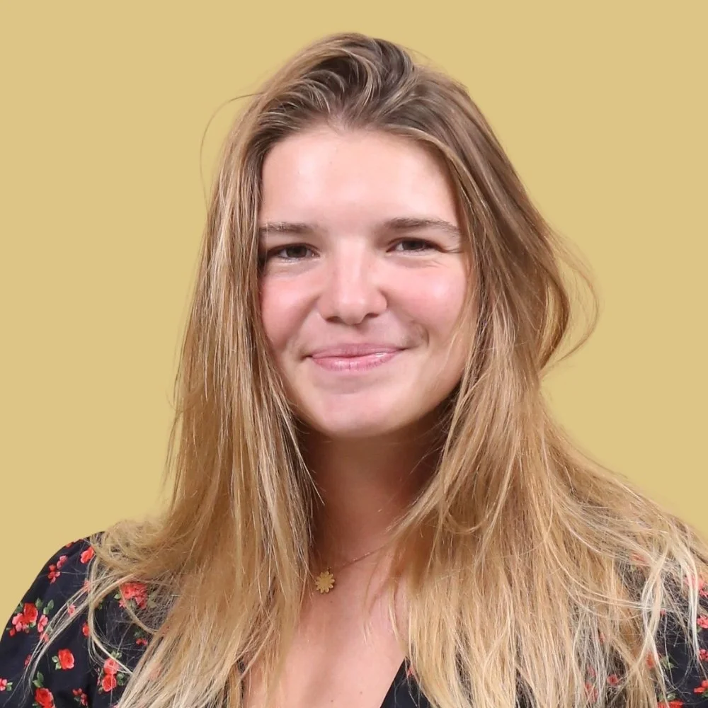 A young woman with long, wavy blonde hair, wearing a black top with a floral pattern and a daisy-shaped necklace, smiling at the camera against a plain yellow background.