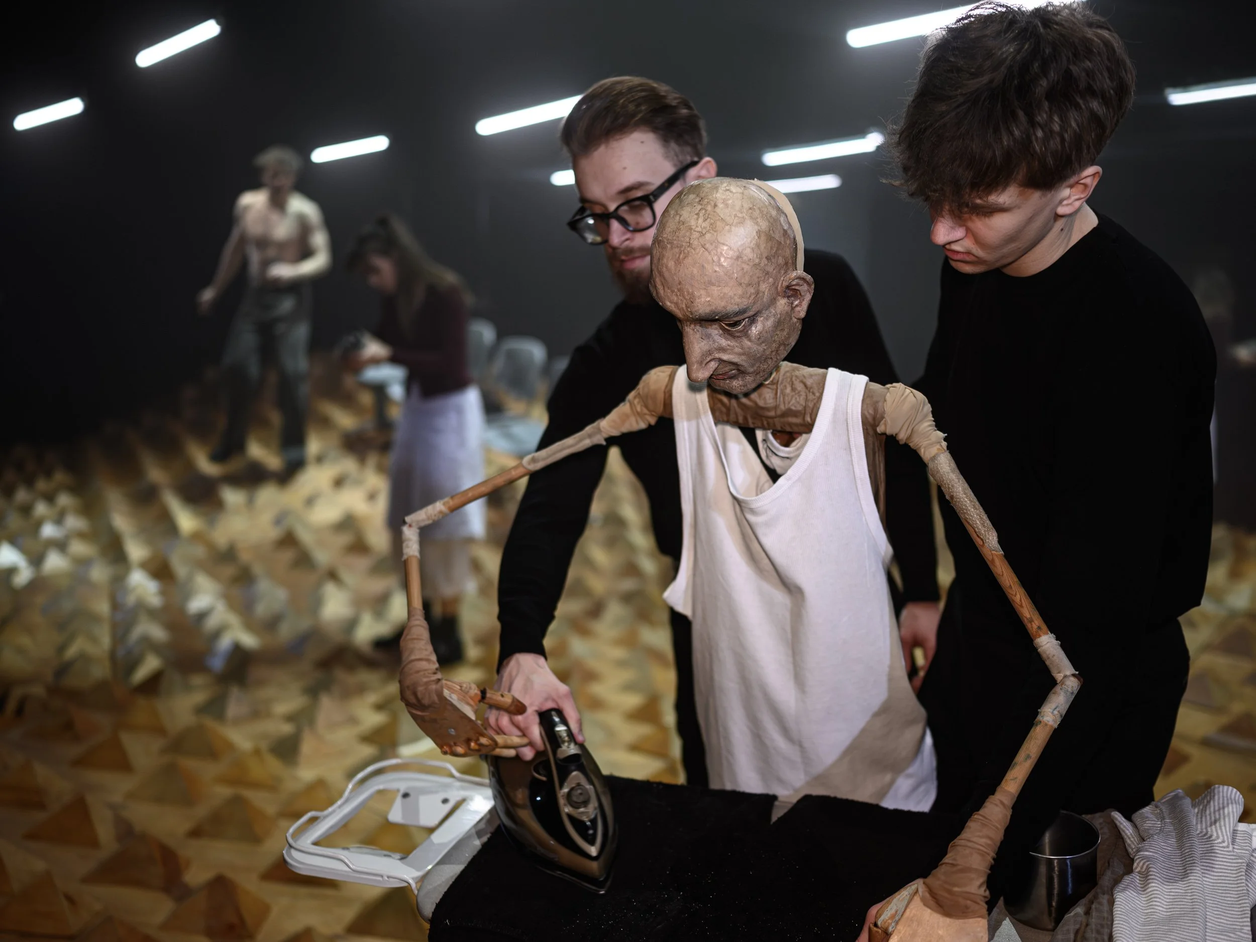 Zwei Menschen arbeiten an einem beleuchteten Kunstprojekt mit einer Puppe, die als lebensechte, alte, gebeugte Figur gestaltet ist, mit einem weißen Kleidungsstück, das auf einem schwarzen Tisch liegt, während im Hintergrund weitere Künstler an ähnli