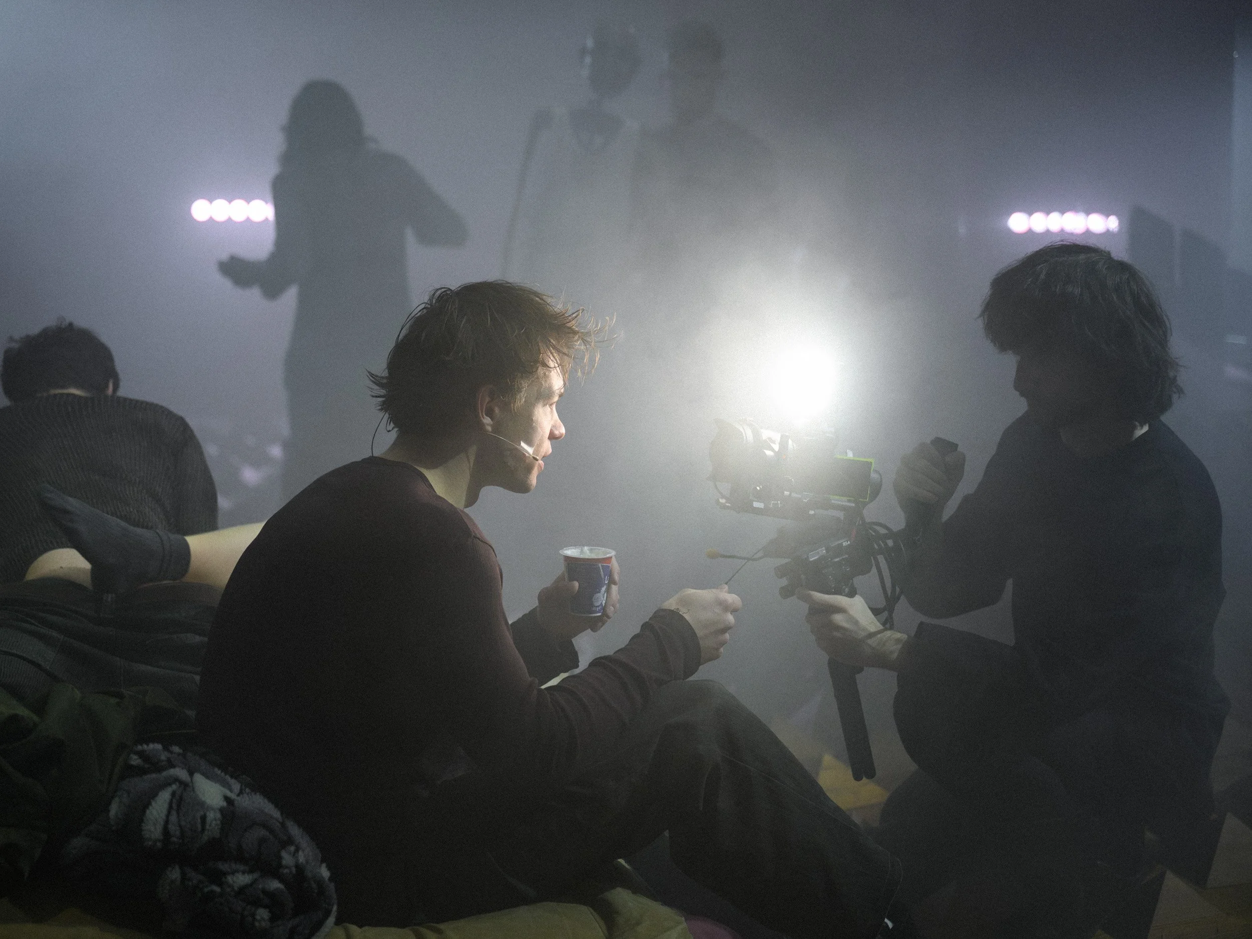 Zwei Personen sitzen nebeneinander, während eine weitere Person im Hintergrund eine Kamera bedient. Es herrscht gedämpfte Beleuchtung mit bunten Lichtern, die eine Veranstaltung wie eine Konferenz oder einen Filmset andeuten.