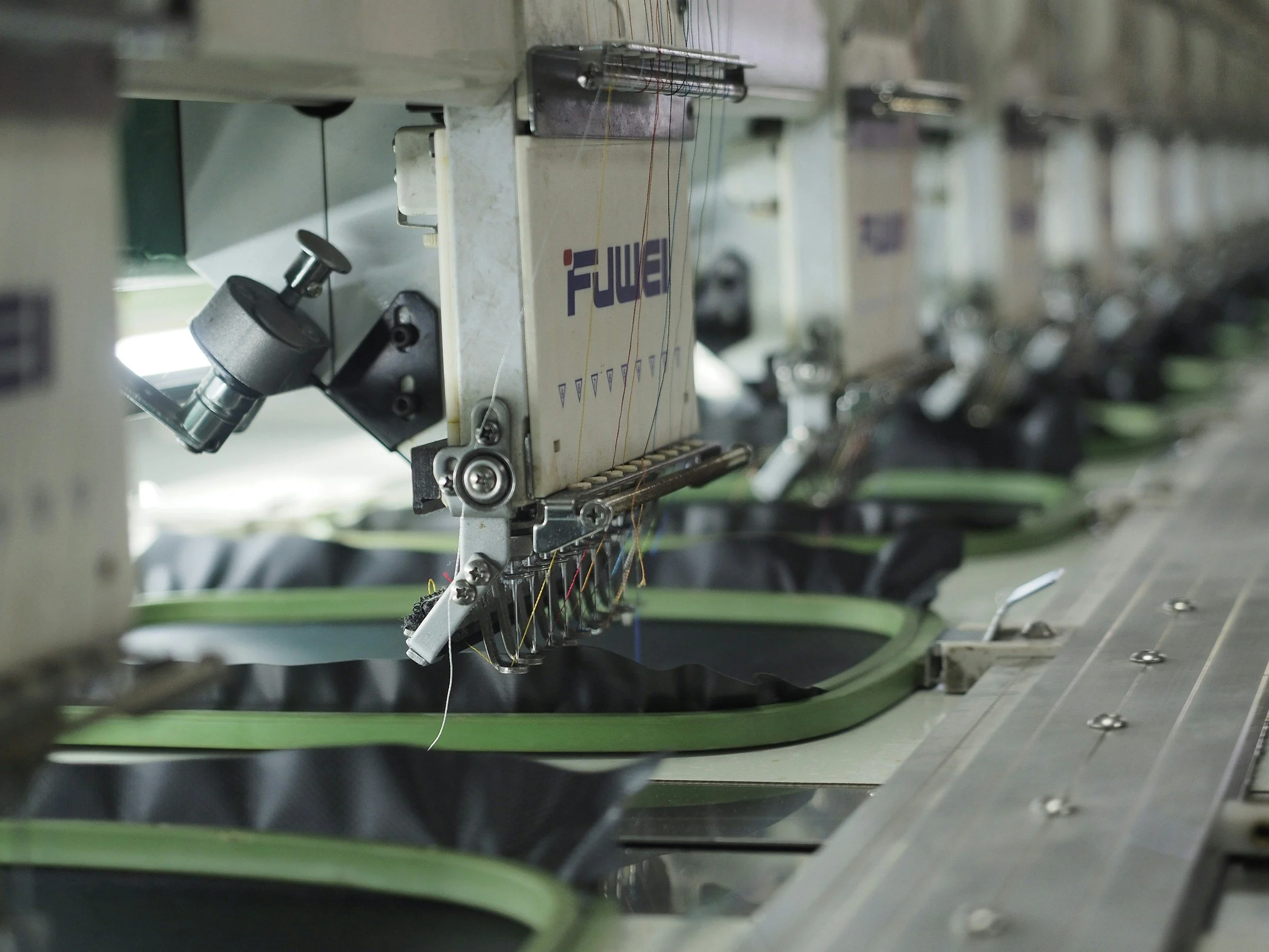 Close-up of an industrial sewing machine stitching shoe uppers in a factory, with multiple sewing stations in the background.