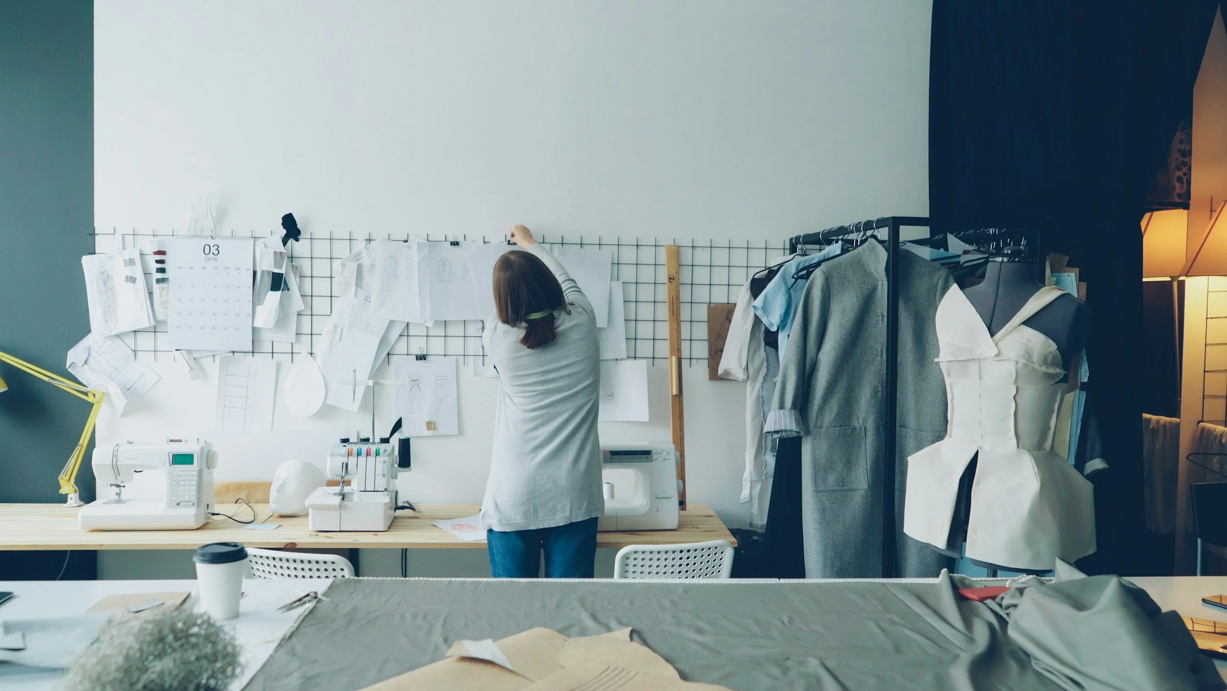 A woman working in a fashion design studio, adjusting fabric sketches on a wire grid wall, surrounded by sewing equipment, fabric, and mannequins with garment prototypes.