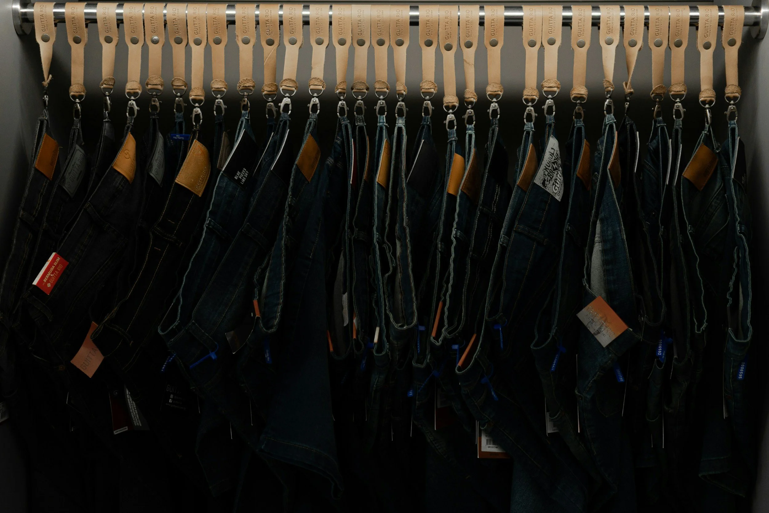 A row of jeans hanging on a clothing rack with beige leather belts at the top.