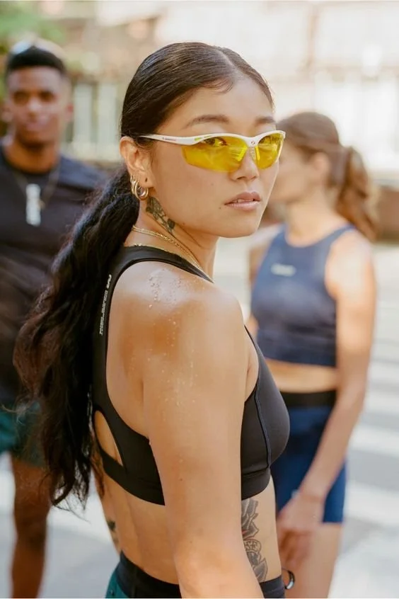 A woman with long dark hair wearing yellow sports sunglasses, a black athletic top, and tattoos on her side, looking over her shoulder during an outdoor activity with other people in the background.