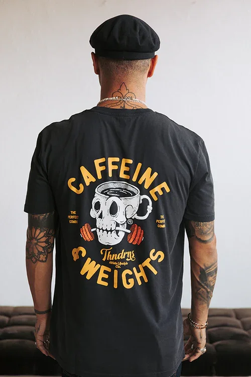 Person wearing a black T-shirt with a skull and coffee cup graphic and the words "Caffeine & Weights" on the back, also has tattoos on arms and neck, and is wearing a black beret.