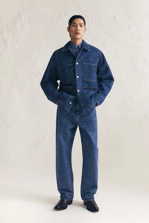 A person standing against a plain white wall, wearing a dark blue denim jacket, matching denim jeans, black shoes, and a dark blue shirt underneath.
