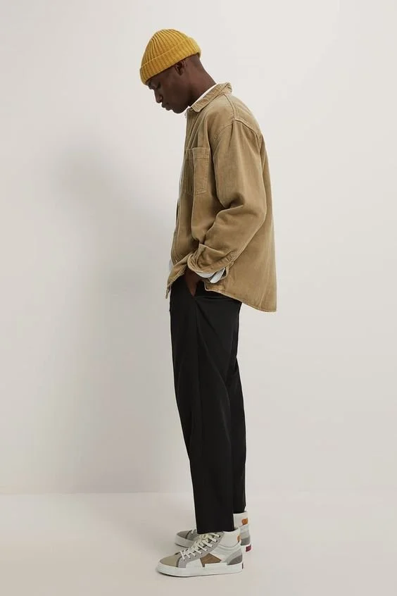 A young man in a beige jacket, yellow knit cap, black pants, and white sneakers standing sideways with his head down and hands in pockets.