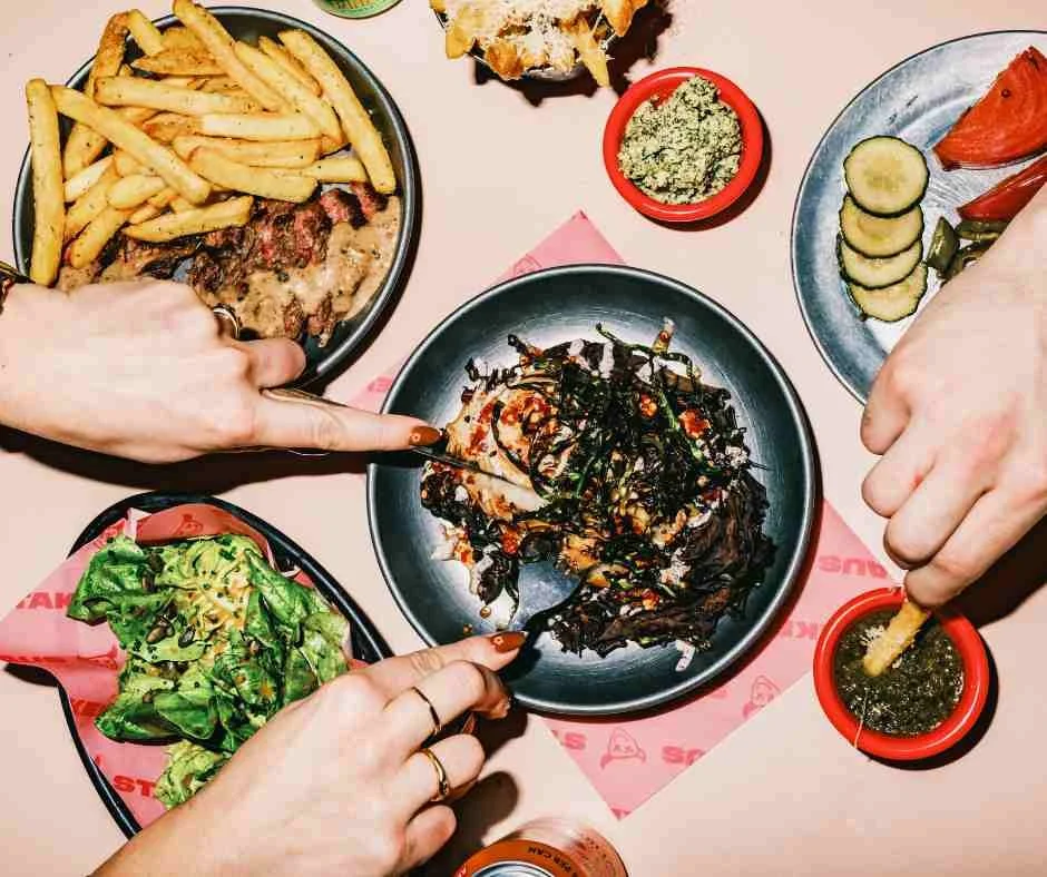 hands over a mix of dishes of chips, salad and steak on a pink table