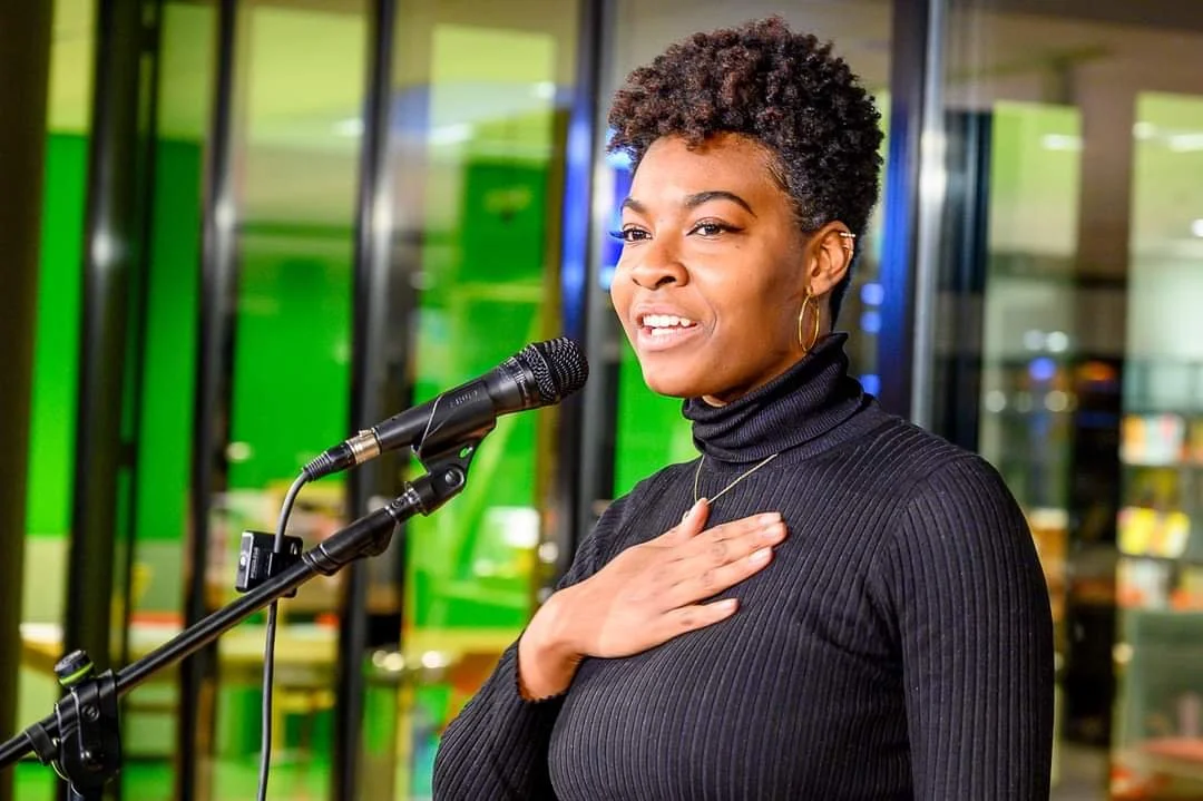A woman with short curly hair giving a speech at a microphone, with her right hand over her chest, wearing a black turtleneck and gold jewelry.