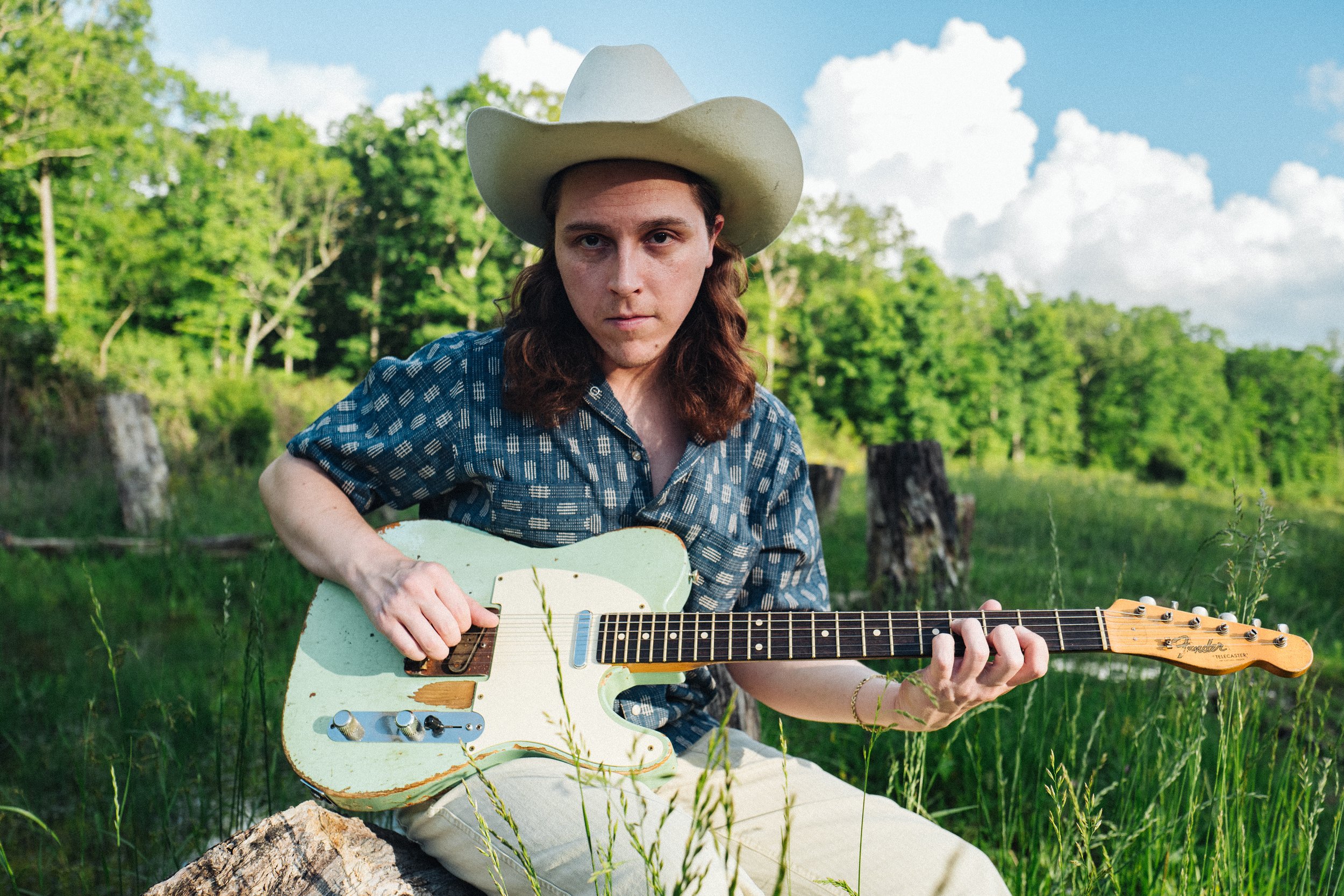 Daniel Donato wearing a cowboy hat playing an electric guitar outdoors in a green, wooded area on a sunny day.