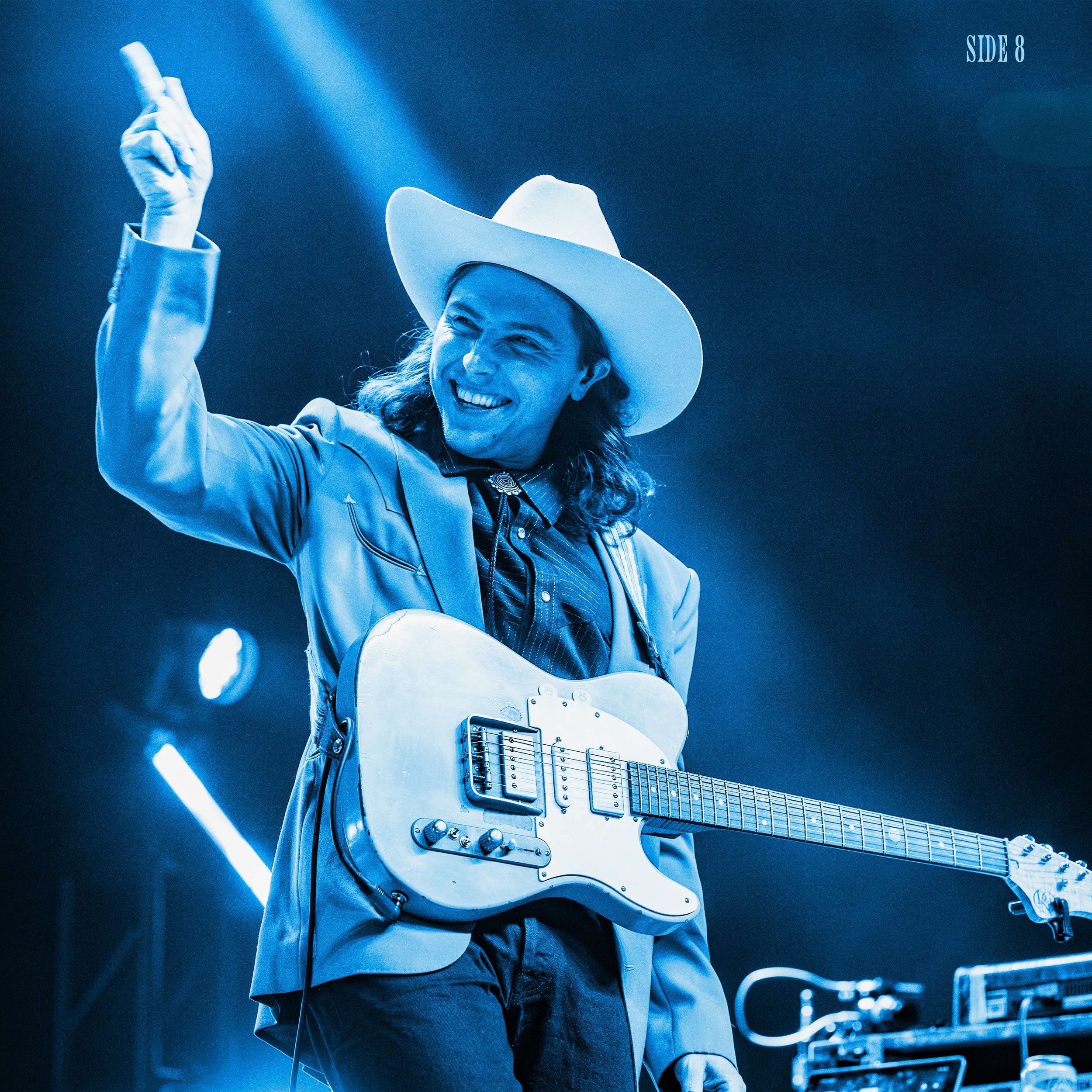 Daniel Donato in a cowboy hat playing an electric guitar on stage, pointing upwards with his right hand, smiling, with stage lights in the background.