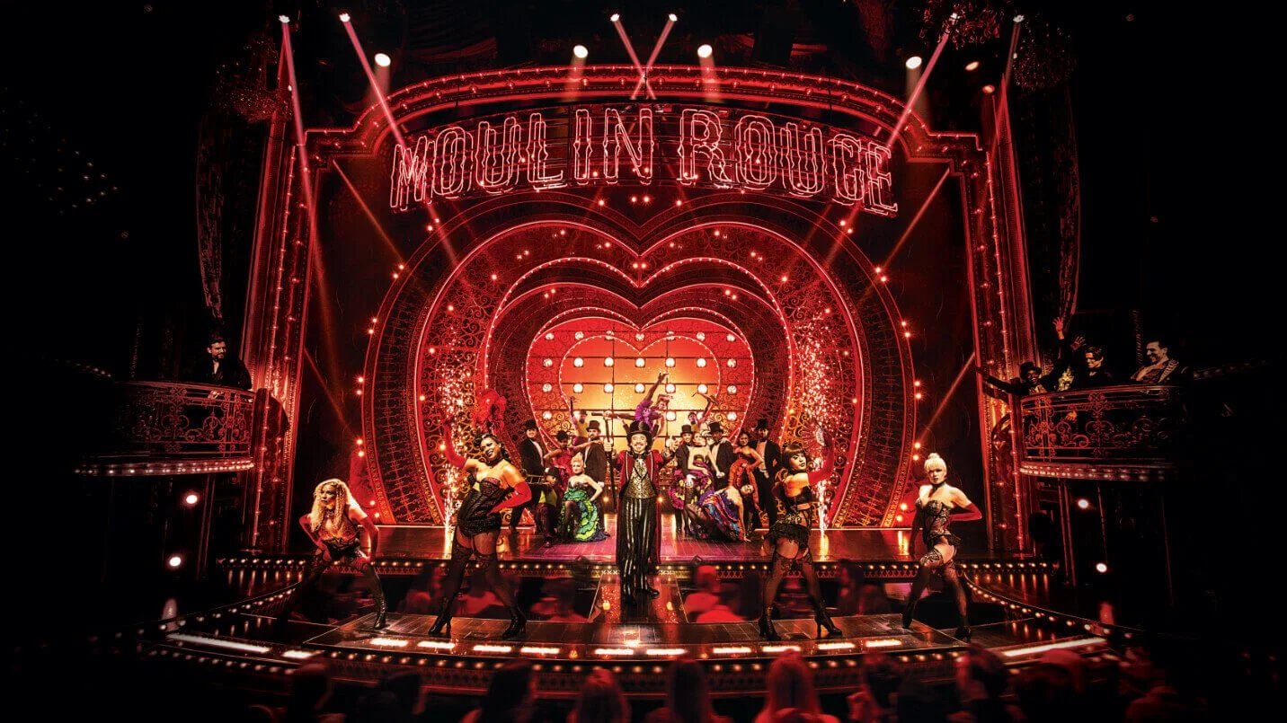 The stage of Moulin Rouge with performers in colorful costumes, a large heart-shaped backdrop, and bright red neon lights spelling out 'MOULIN ROUGE' at the top.