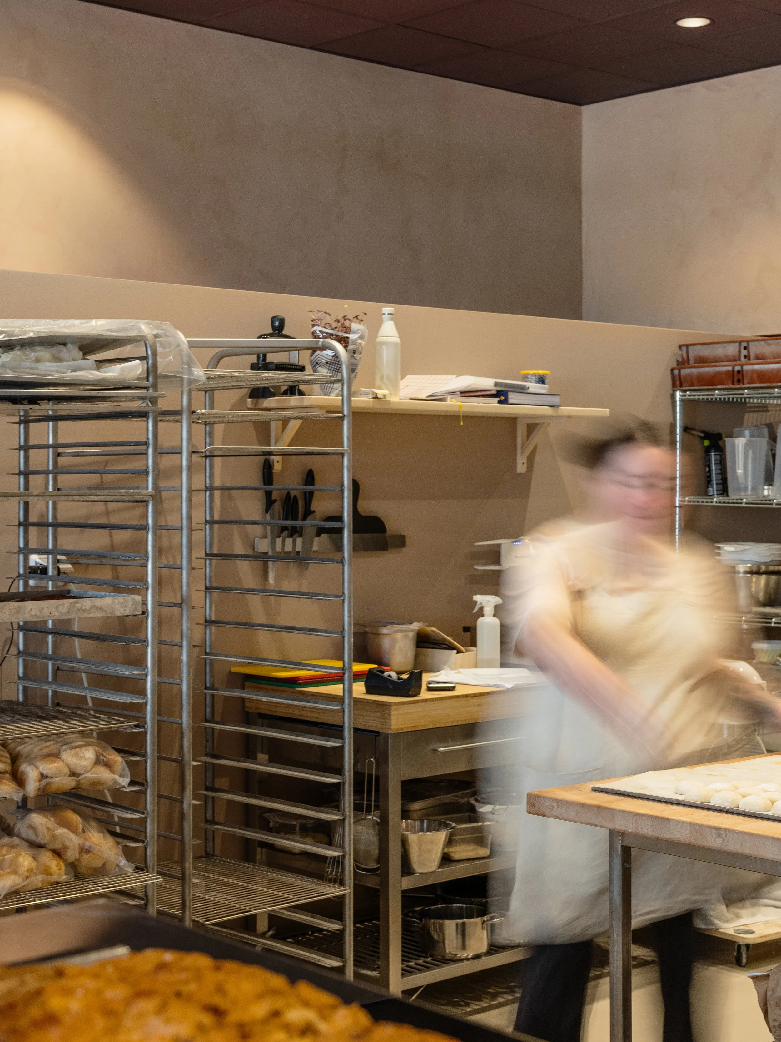 Work area in an open bakery with baking racks, worktables and ongoing food preparation.