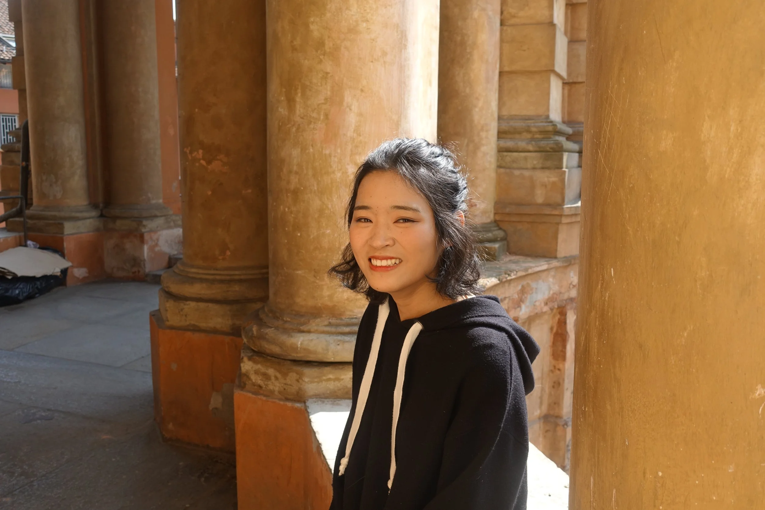 Young woman smiling outdoors near old colorful columns and stone steps, wearing a black hoodie.