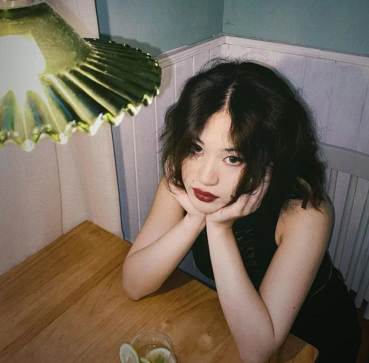 A woman with dark, wavy hair and red lipstick sitting at a wooden table, resting her face in her hands, with a glass of drink in front of her, and a green lamp partially visible in the top left corner.