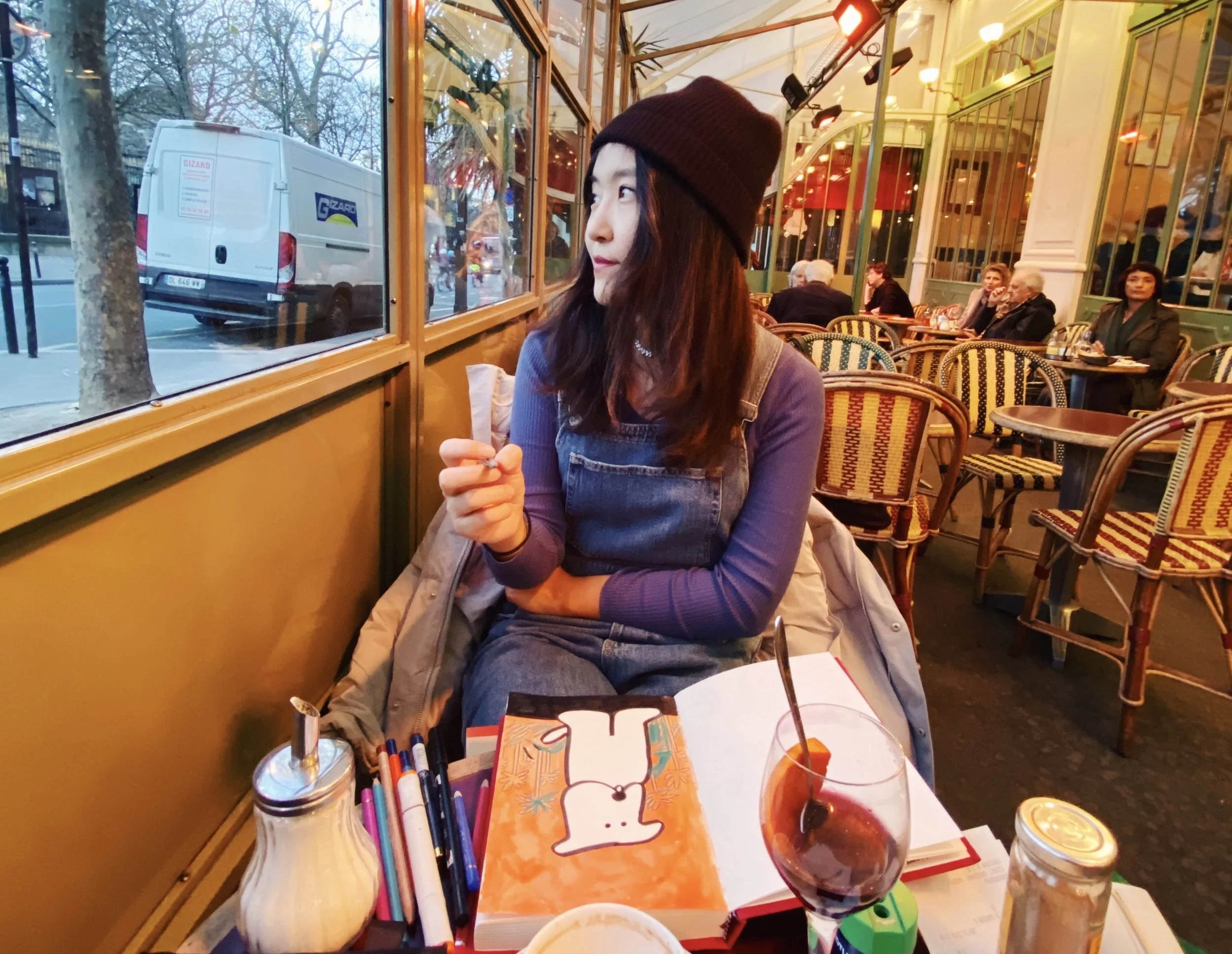 A woman wearing a black beanie and denim overalls sitting in a colorful indoor cafe, looking out the window at a street scene with parked trucks and trees.