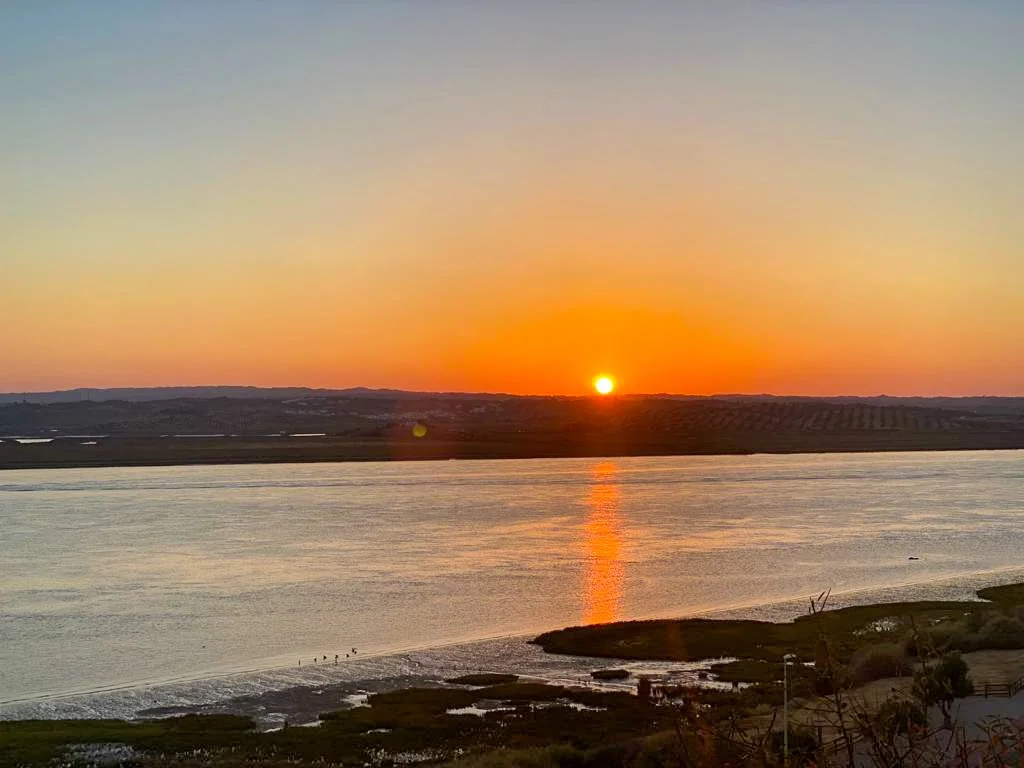 Sonnenuntergang über einem Fluss mit Sonnenreflexion auf dem Wasser, umgeben von Landflächen und einem bewölkten Himmel.