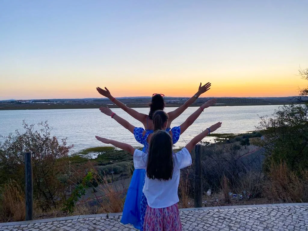Drei Frauen stehen mit ausgestreckten Armen während des Sonnenuntergangs an einem Flussufer.