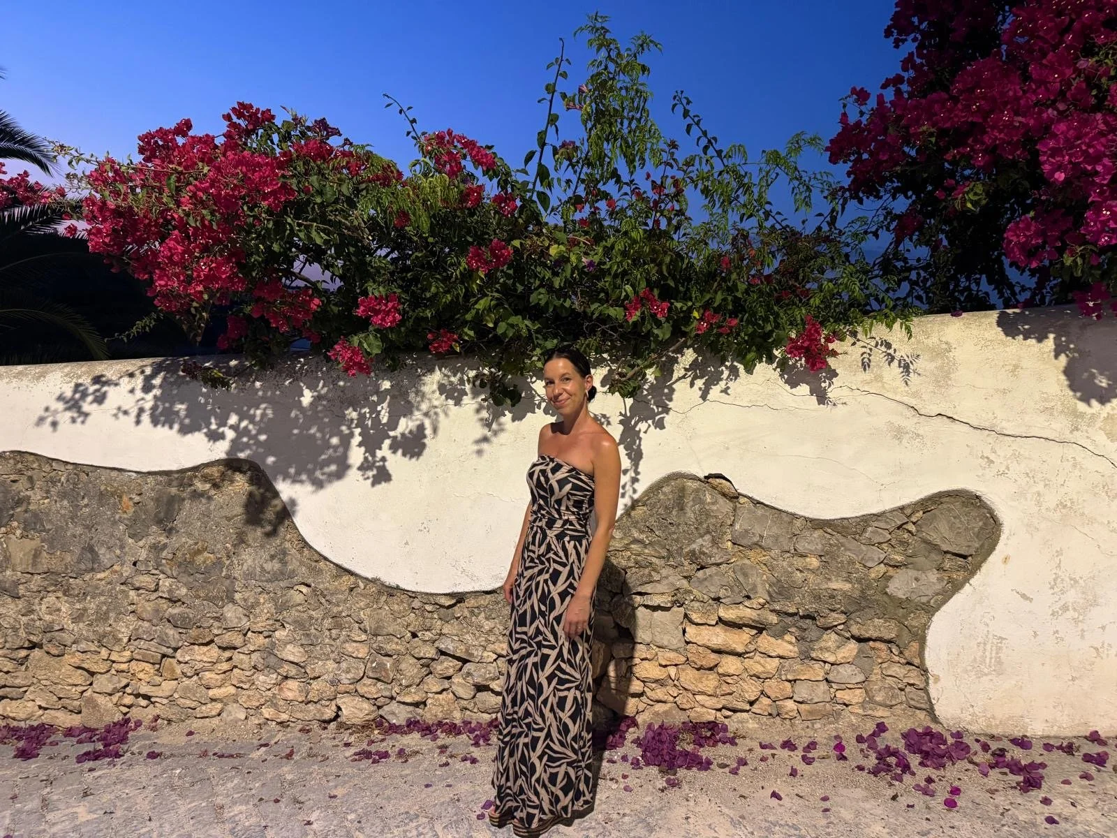 Eine Frau in einem gestreiften, bodenlangen Kleid steht vor einer weißen Mauer mit einer künstlerischen Steinverzierung. Im Hintergrund sind Blumen und Sträucher, die bei Sonnenuntergang pinke Blüten tragen, zu sehen.