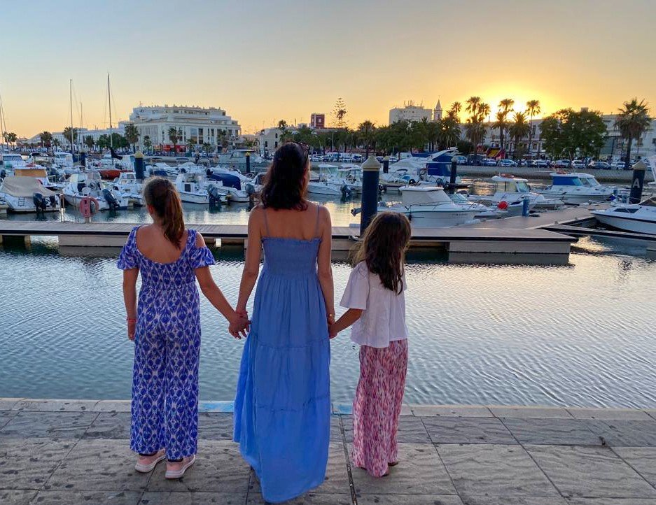 Drei Frauen halten am Hafen bei Sonnenuntergang Händchen, mit Booten und Palmen im Hintergrund.