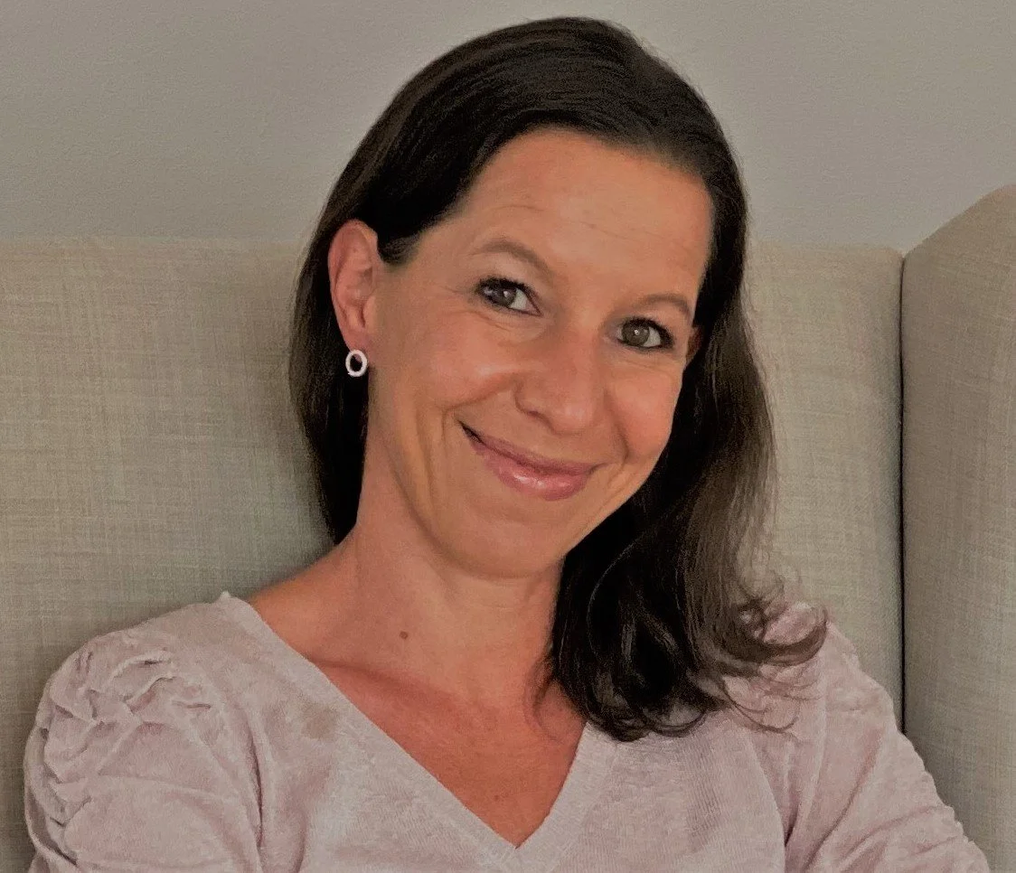 Porträt einer lächelnden Frau mit dunklen, mittellangen Haaren, in einem hellrosa Oberteil, sitzend auf einem beige Sitz.