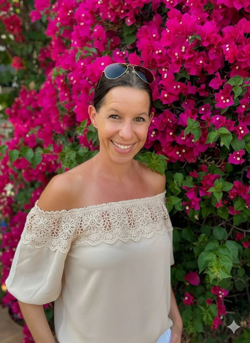 Frau mit Sonnenbrille auf dem Kopf, lächelnd vor einer Bougainvillea-Blumenwand, trägt ein off-Shoulder-Kleid mit Spitzendetail.