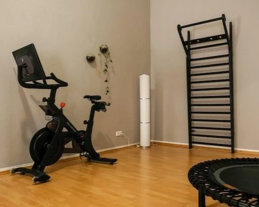 Atemwerk Longevity Zentrum mit Fitnessgeräten, darunter ein Heimtrainer, eine Sprossenwand und ein Minitrampolin. Der Boden ist aus Holz. Es gibt Dekorationen an der Wand.