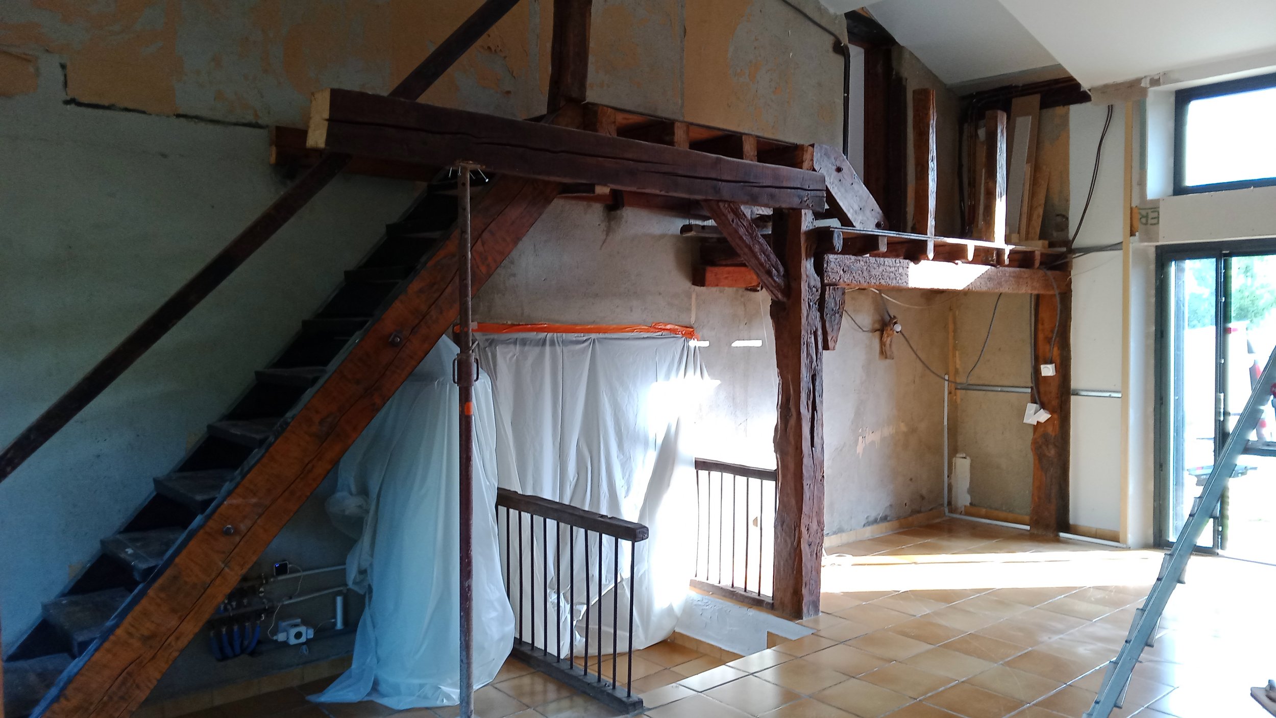 Intérieur en rénovation avec poutres en bois apparentes, escalier en métal, fenêtres laissant entrer la lumière naturelle, et des protections de chantier.