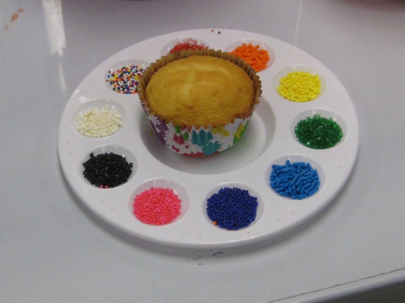 Cupcake on a white circular palette surrounded by various colored sprinkles in small white containers.
