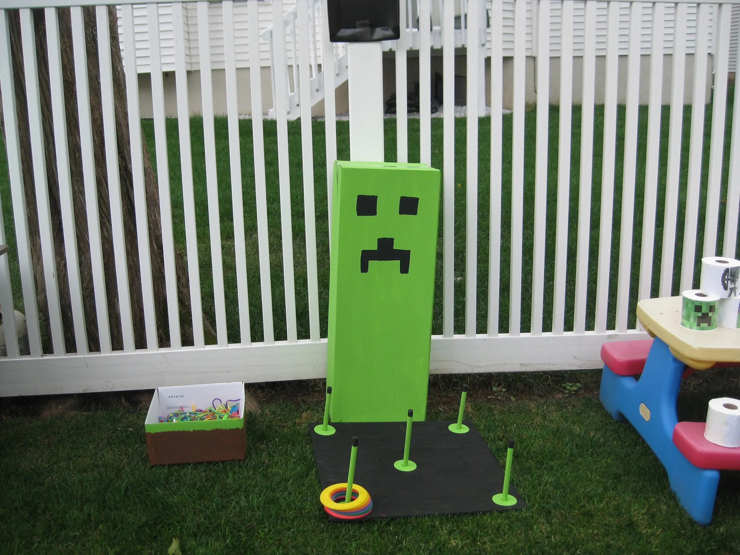 A green Minecraft-themed cornhole board with a pixelated face, set up on a black mat with five green cones on a grassy yard, with a white fence in the background and some colorful toys on a small blue and pink table to the right.