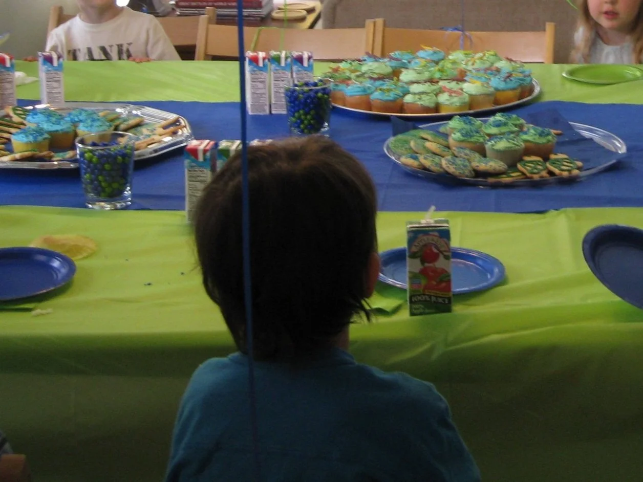Children celebrating a birthday with a table of decorated cupcakes, cookies, juice boxes, and party snacks.